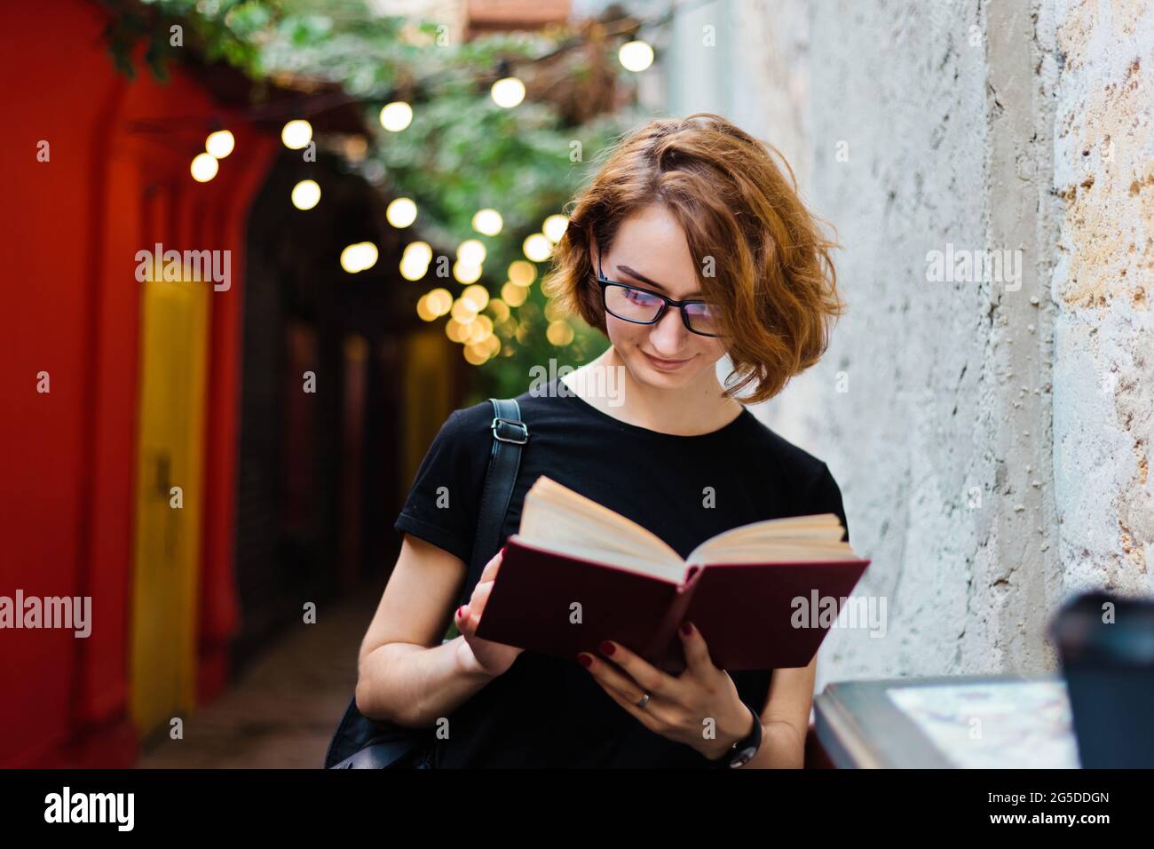 Junge Hipster-Frau in Brille mit kurzen Haarschnitten Lesebuch im Straßencafe im Freien Stockfoto