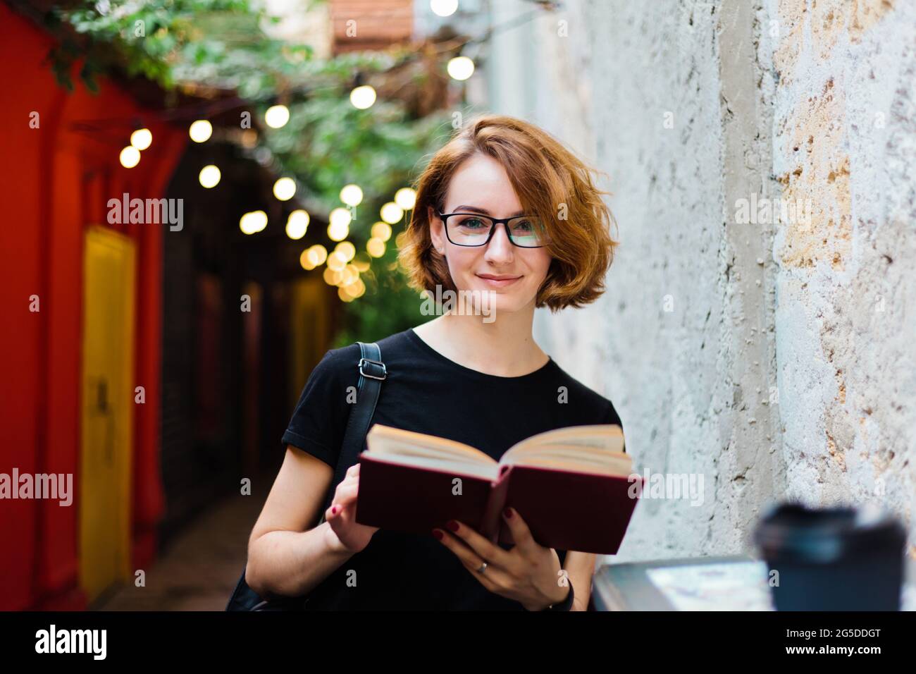 Junge Hipster Frau in Brille mit kurzen Haarschnitten Lesebuch im Straßencafe im Freien und Blick auf die Kamera Stockfoto