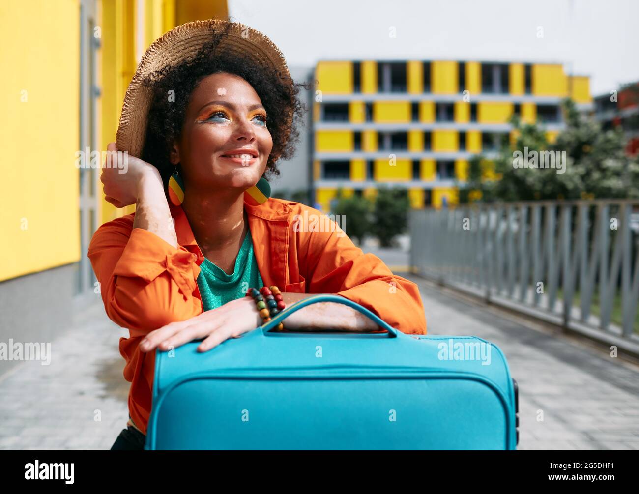 Lustige multiethnische Frau mit hellem Make-up und farbigen Kleidern mit einem Koffer reist durch schöne urbane Orte Stockfoto