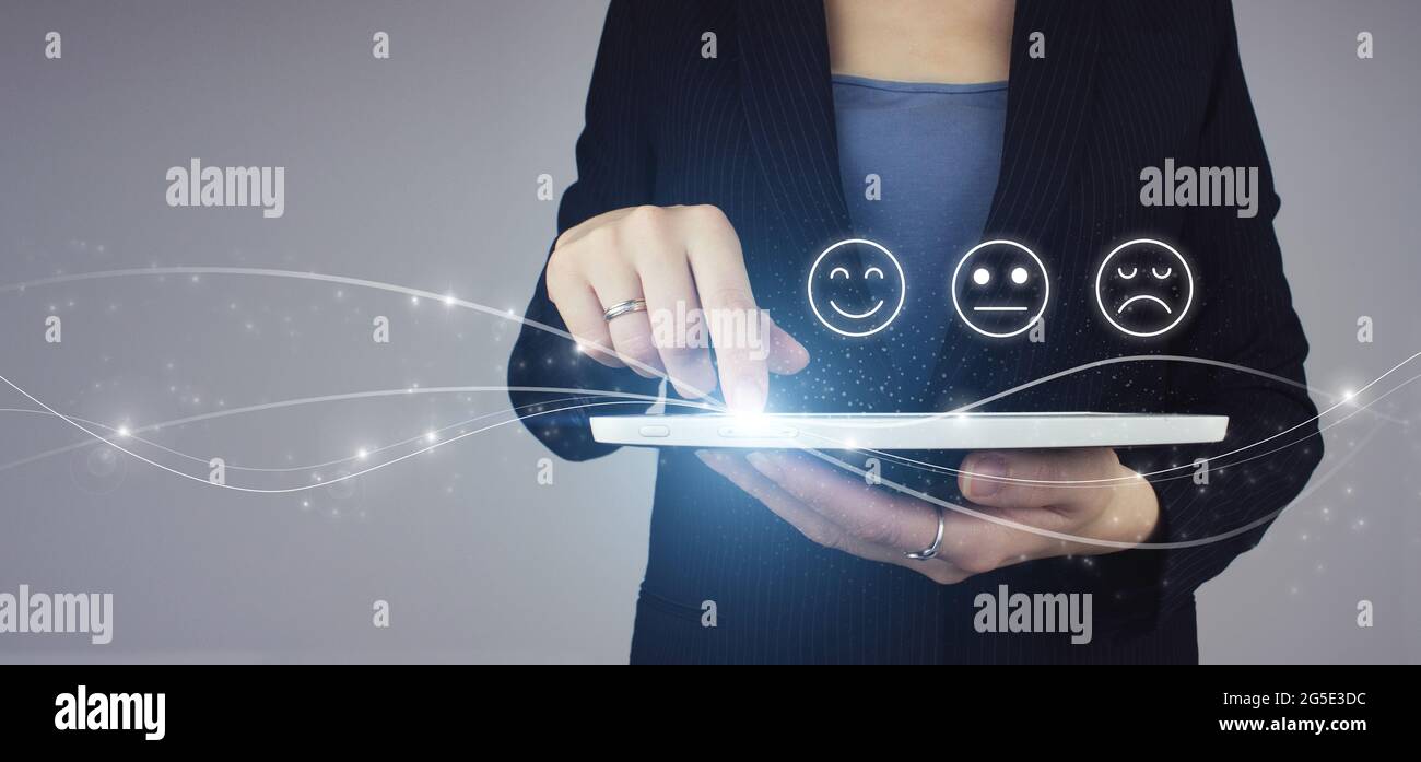 Smiley Face Rating für eine Zufriedenheitsumfrage. Weißes Tablet in Geschäftsfrau Hand mit digitalem Hologramm Gesicht Emotion Zeichen auf grauem Hintergrund. Kunde Stockfoto