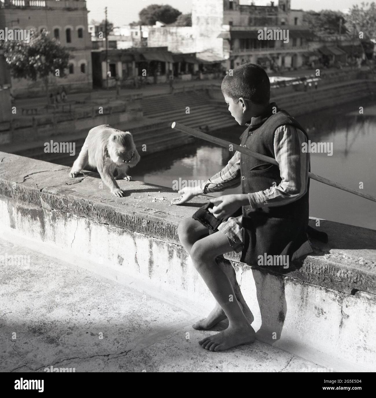 In den 1950er Jahren, historisch, Benares, Indien, sitzt auf einer Steinbrücke, ein junger barfüßiger Junge, mit einem Holzstab unter seinem Arm, der einen Straßenaffen füttert. Stockfoto