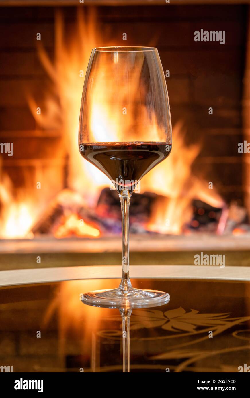 Gemütliche Kamin-Szene und ein Glas Rotwein, Spiegelung von einem Feuer auf einem Glastisch. Stockfoto