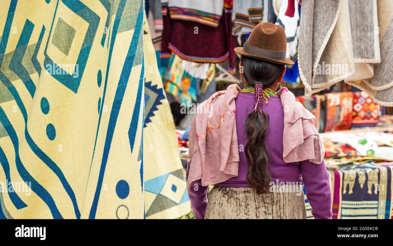 Indigene ecuadorianische Otavalo Frau in traditioneller Kleidung, Hut und Frisur auf dem lokalen Markt von Otavalo mit Textil- und Stoffständen, Ecuador. Stockfoto