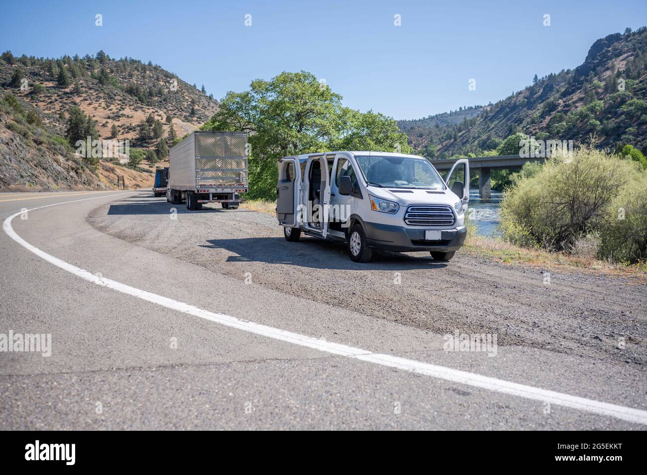 Kommerzieller kompakter Mini-Van und beladene große LKW-Sattelschlepper mit Sattelaufliegern machen eine Pause auf der Straßenschulter am Ufer eines Berg-riv Stockfoto