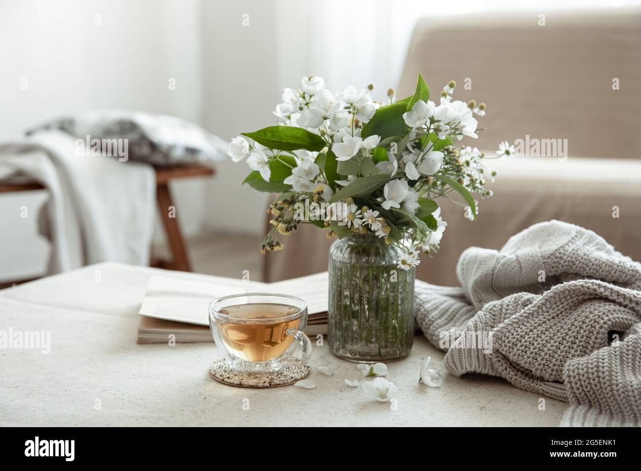 Stillleben mit einer Tasse Kräutertee, einem Blumenstrauß, einem Buch und einem gestrickten Element auf einem verschwommenen Hintergrund. Stockfoto