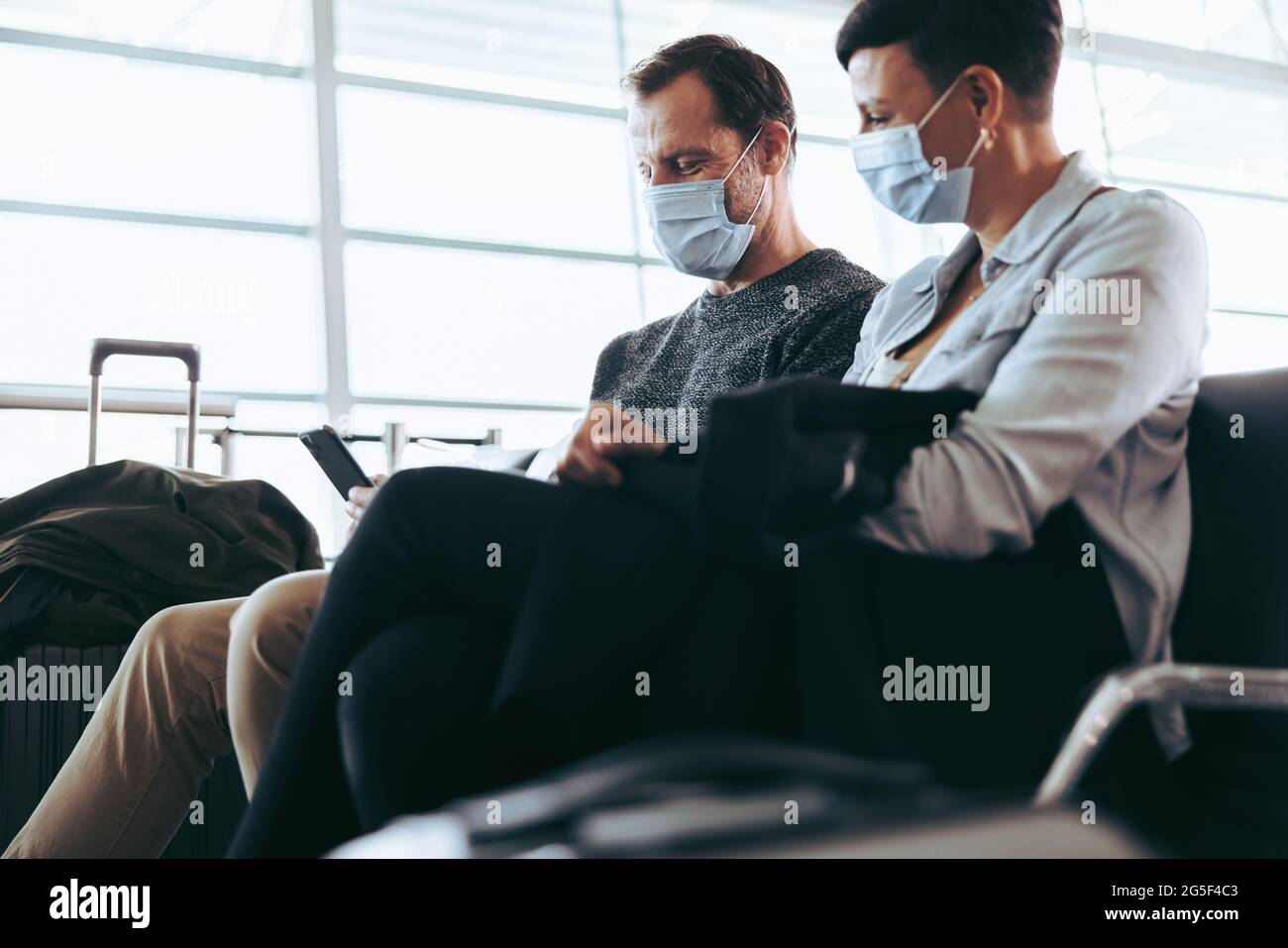 Mann überprüft während einer Pandemie das Handy, während eine Frau am Abflugbereich des Flughafens sitzt. Paar sitzt am Flughafen mit Gesichtsmaske und Smartphone. Stockfoto
