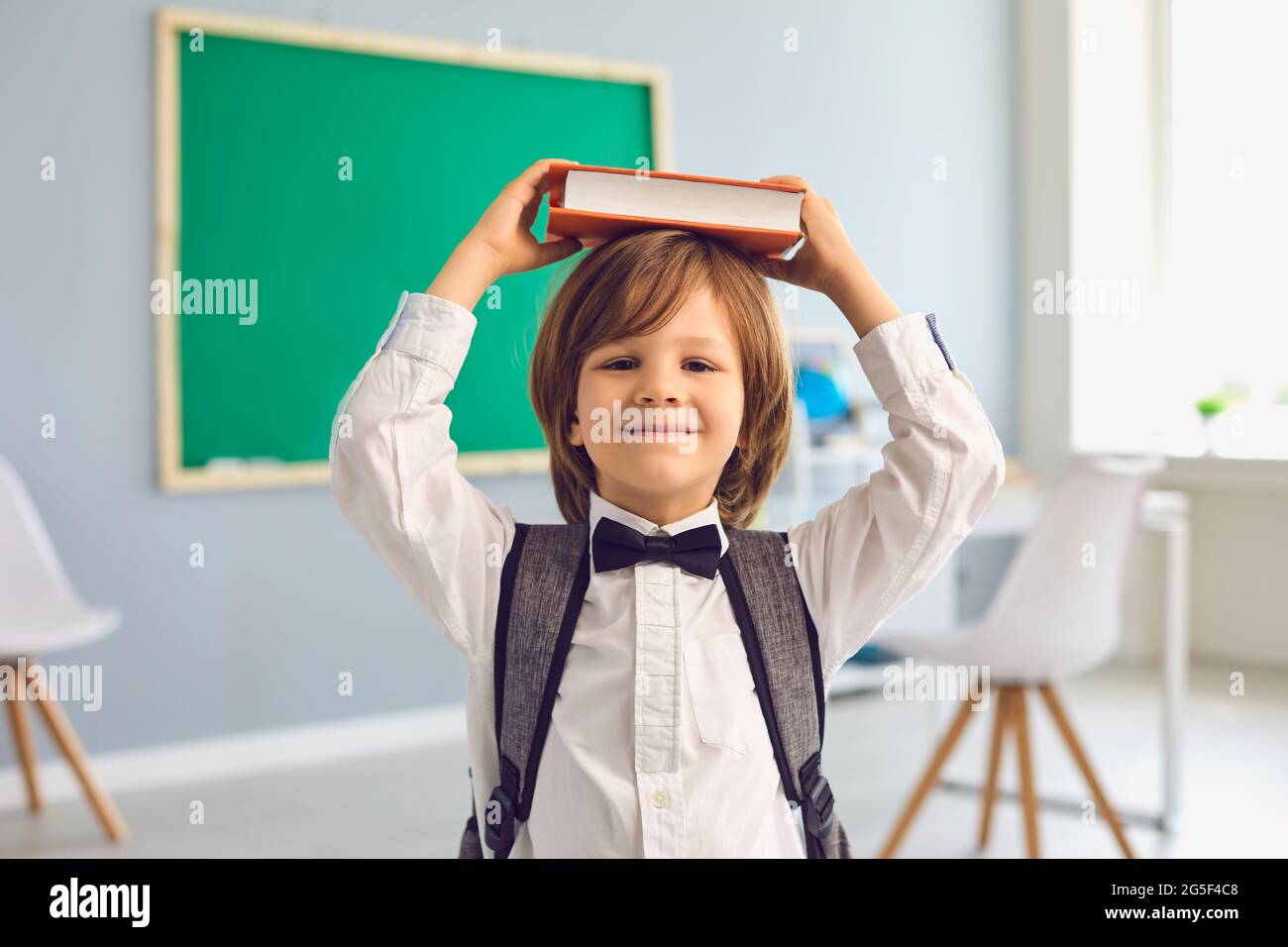 Schulbildung. Zurück zur Schule. Ein lustiger Schuljunge hält ein Buch über seinen Kopf, während er in der Schule auf einem Schreibtisch sitzt Stockfoto