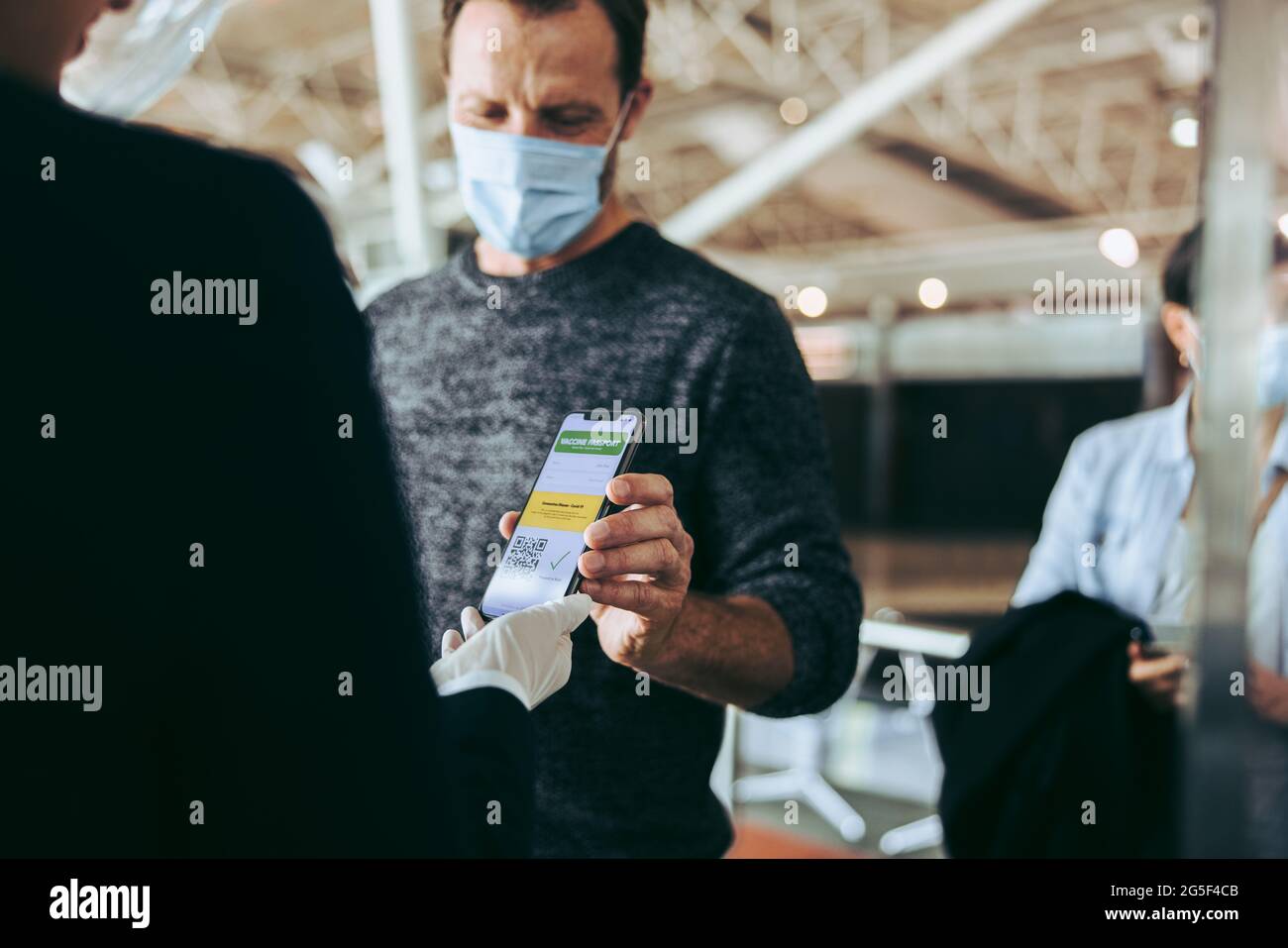 Mann in Gesichtsmaske, der seinen covid-19-Impfpass auf dem Handy zeigt. Der Reisende am Check-in-Schalter am Flughafen zeigt seine impfserie Stockfoto