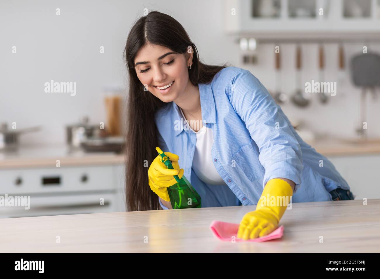 Fröhliche lässige junge Frau in gelben Gummihandschuhen Reinigungstisch in gemütlicher weißer Küche, mit Spray und Staubtuch. Selektiver Fokus, kostenlose Kopie Spa Stockfoto