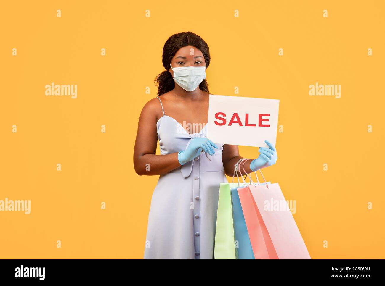 Pandemisches Einkaufen. Millennial schwarze Dame in Schutzmaske und Handschuhen zeigt VERKAUFSSCHILD an der Kamera, hält Einkaufstaschen, Kauf von Waren auf Discount Dur Stockfoto