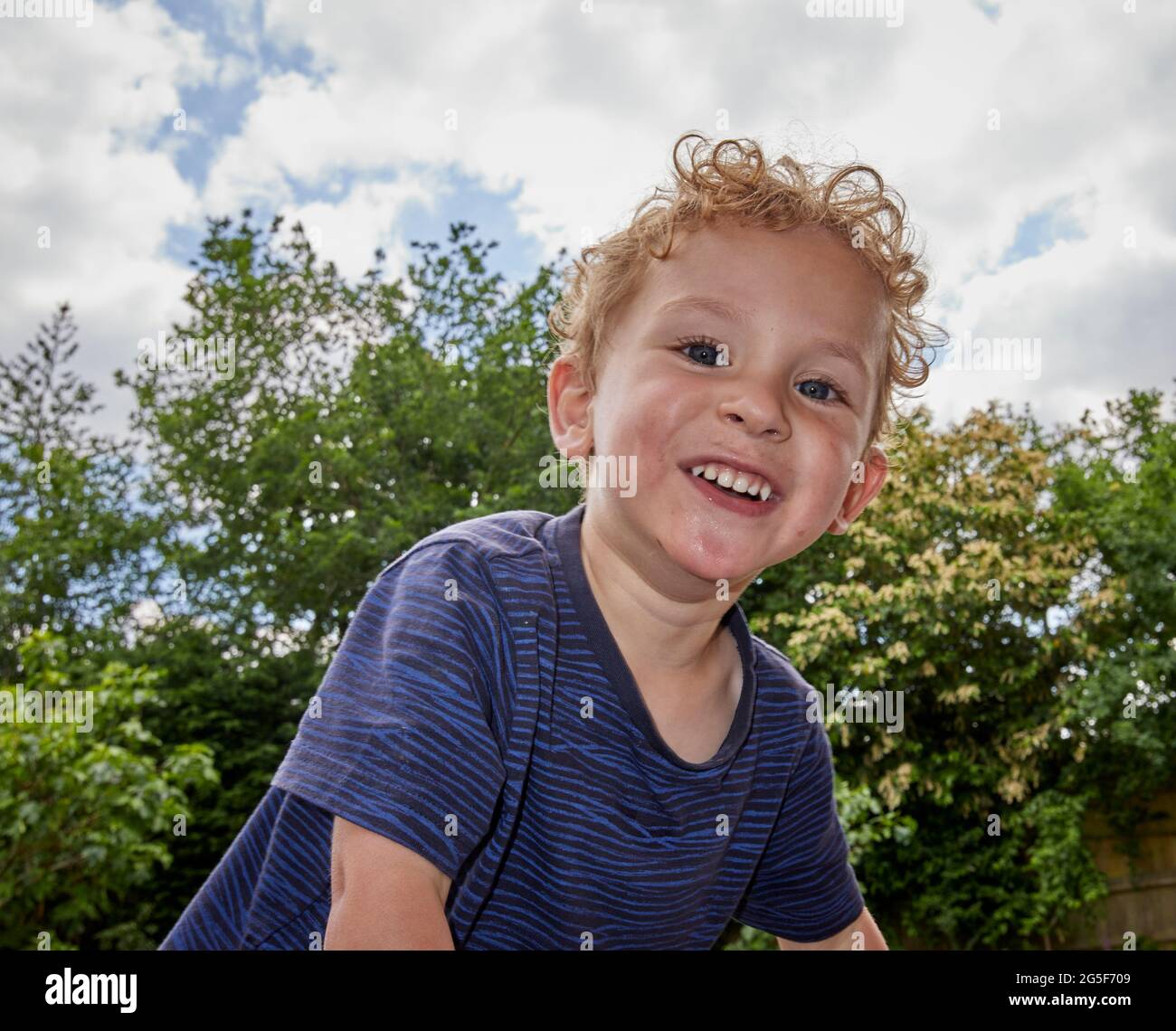 Ein lächelnder, fröhlicher weißer kaukasischer kleiner Junge im Alter von fast 3 Jahren mit lockigen blonden Haaren in einem Garten, der ein blaues Oberteil trägt, lächelt an einem sonnigen Tag in en an der Kamera Stockfoto