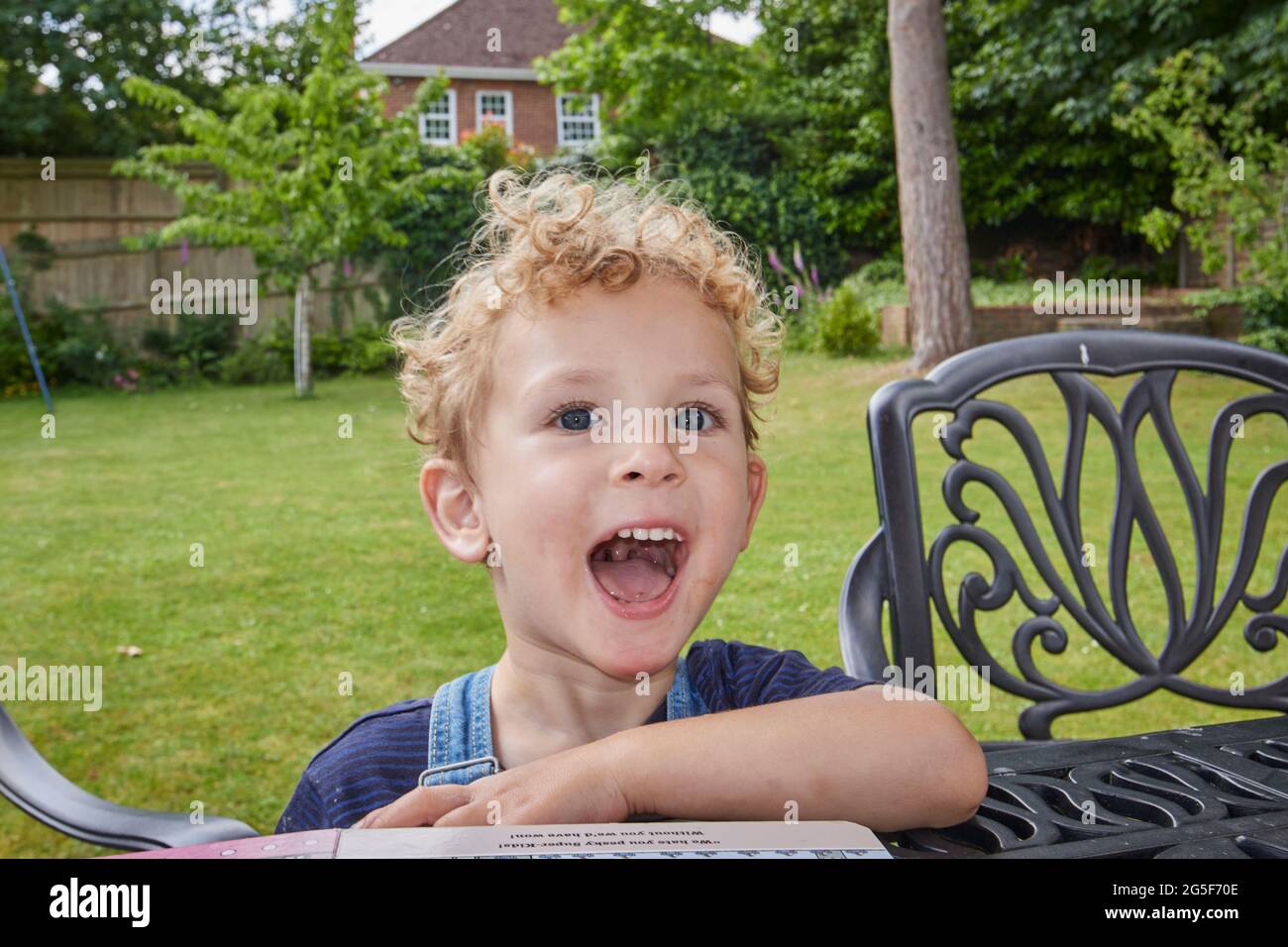 Ein glücklicher weißer kaukasischer kleiner Junge im Alter von fast 3 Jahren mit lockig blonden Haaren sitzt an einem Tisch in einem Garten an einem sonnigen Tag in England mit offenem Mund Stockfoto