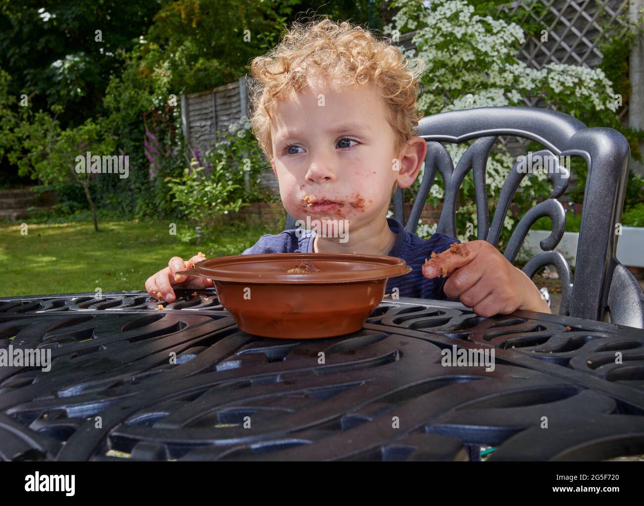 Ein klebriger weißer kaukasischer kleiner Junge im Alter von fast 3 Jahren mit lockigem blondem Haar sitzt im Freien mit einem schmuddeligen braunen Mund, der Schokoladenkuchen isst Stockfoto