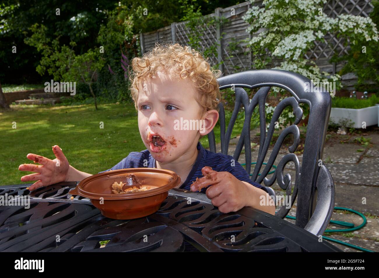 Ein klebriger weißer kaukasischer kleiner Junge im Alter von fast 3 Jahren mit lockigem blondem Haar sitzt im Freien mit einem schmuddeligen braunen, offenen Mund, der Schokoladenkuchen isst Stockfoto