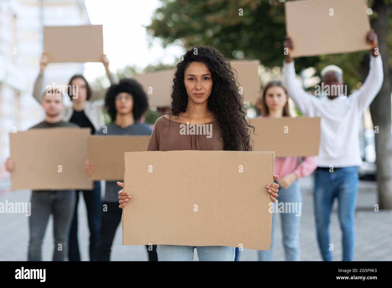 Aktive, hübsche junge Frau, die eine Gruppe von Demonstranten auf der Straße führt. Internationale, vielfältige Gruppe von Menschen mit leeren Plakaten, die gegen Brummen protestierten Stockfoto