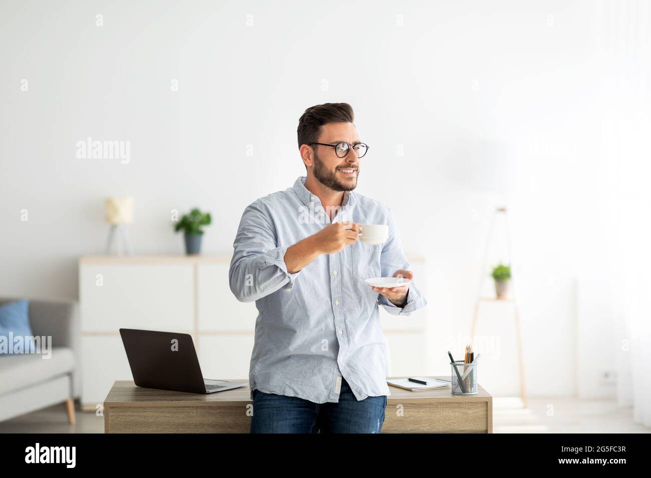 Junger Freiberufler mit einer Tasse Kaffee, der am Schreibtisch in der Inneneinrichtung stand, sich am Tisch lehnte und wegschaute, heiße Morgengetränke genoss und Platz zum Kopieren Stockfoto