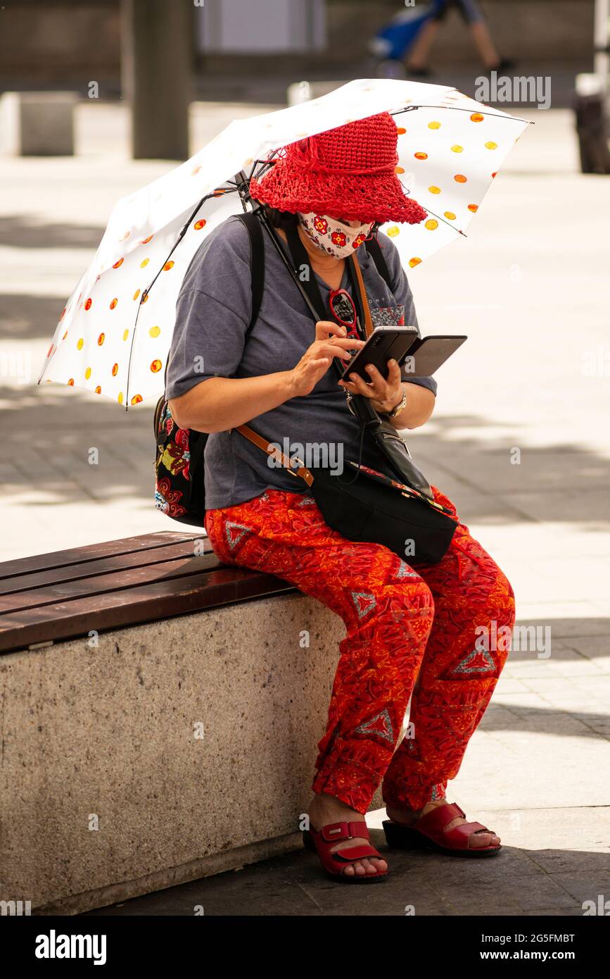 Alleinreisende ältere Frau, die Gesichtsmaske, bunte Tücher und Regenschirm mit dem Smartphone trägt, während sie auf einer Bank im städtischen Umfeld sitzt Stockfoto