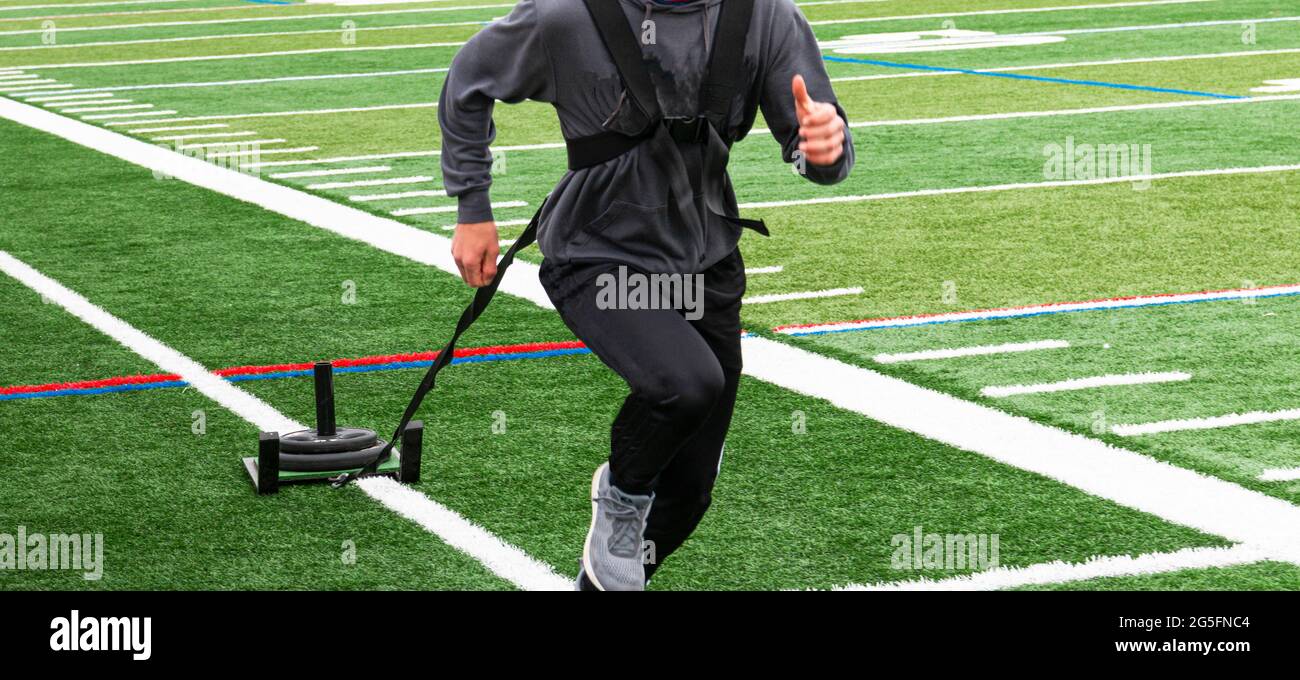 Nahaufnahme os ein Athlet in einem Trainingsanzug zieht einen Schlitten mit Gewichten auf einem grünen Rasenfeld während des Trainings auf der Strecke und im Feld. Stockfoto