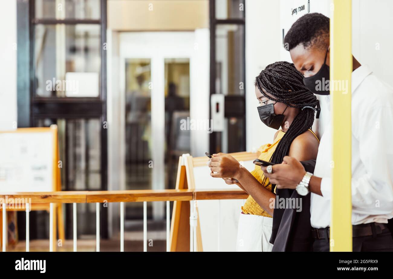 Zwei afrikanische Studenten mit Maske, die ihre Handys auf dem Campus benutzen Stockfoto