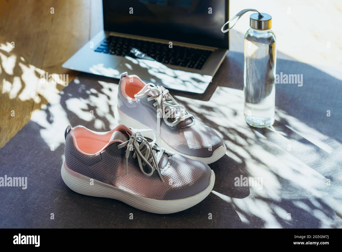Laptop, Sneaker und Wasserflasche auf grauem Yogamatte-Hintergrund mit direkter Sonneneinstrahlung und Schatten. Online-Fitness-Programm, Home Workout. Workout Stockfoto