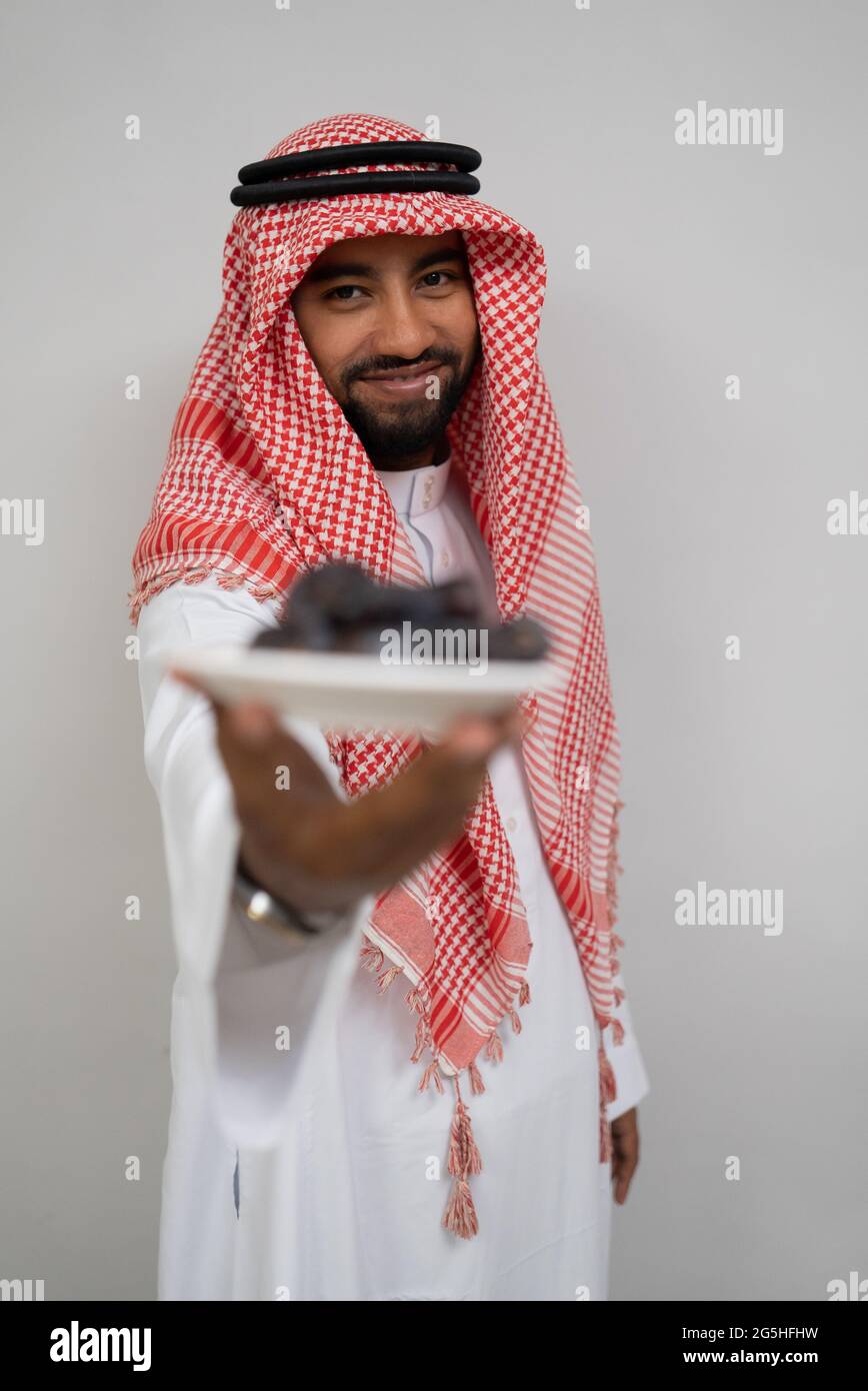 Ein araber im Turban lächelt beim Tragen und bietet der Kamera einen Teller mit Datteln an Stockfoto