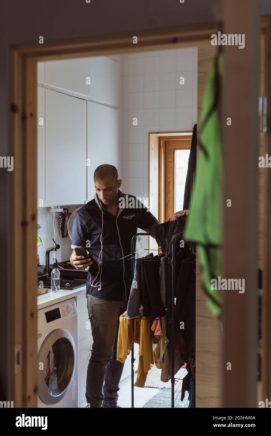 Mann, der Smartphone benutzt, während er im Hauswirtschaftsraum seine Wäsche wäschet Stockfoto
