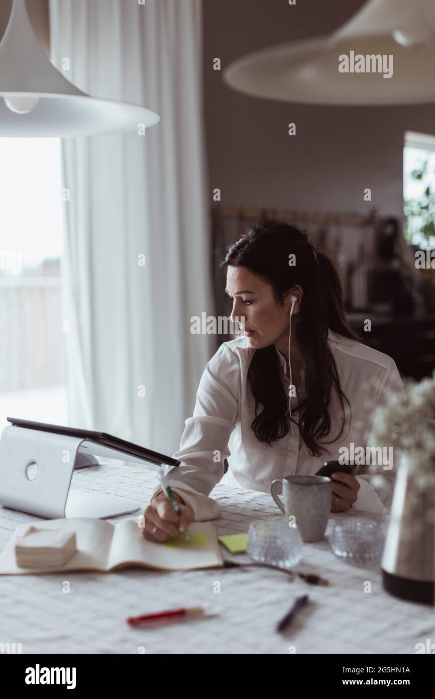 Geschäftsfrau, die im Tagebuch schreibt, während sie zu Hause ein digitales Tablet benutzt Stockfoto