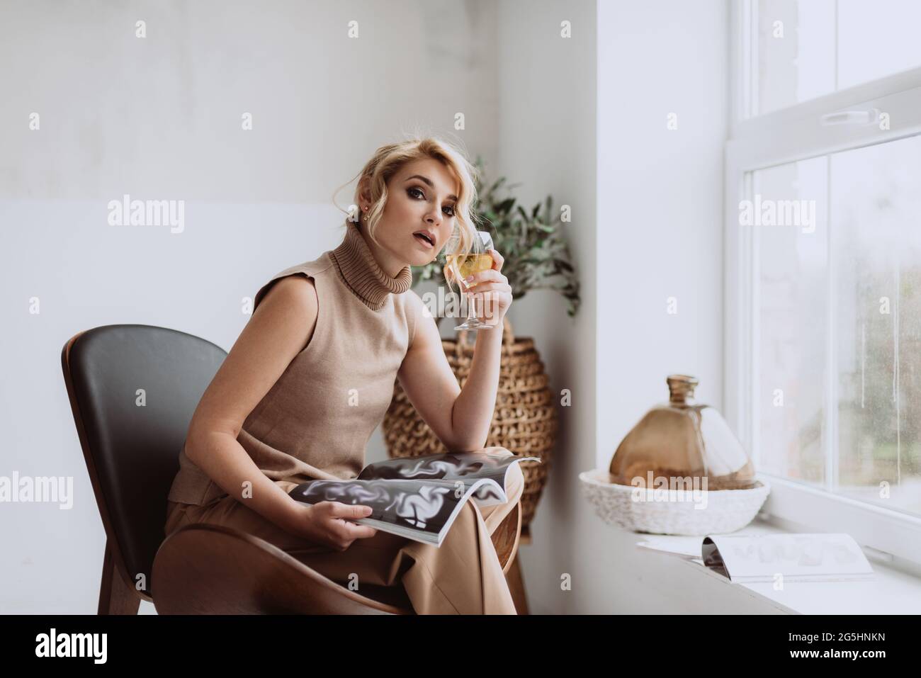 Junge Geschäftsfrau arbeitet zu Hause. Das nachdenkliche, ruhige Model hält ein Glas Weißwein und eine Zeitschrift und blickt auf die Kamera. Schöne Frau in einem modernen Stockfoto