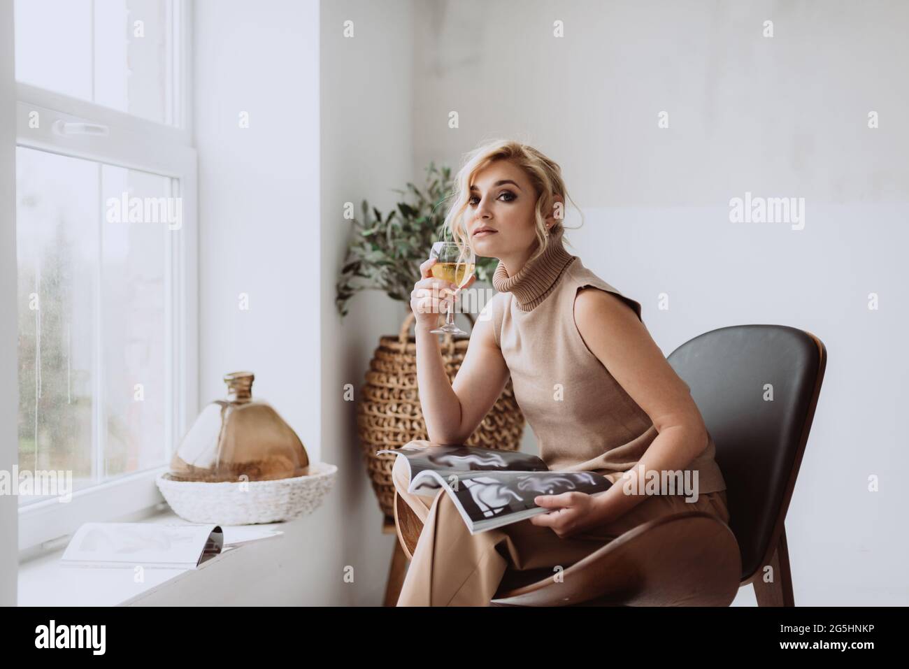 Die junge Geschäftsfrau arbeitet zu Hause.das nachdenkliche, ruhige Model hält ein Glas Weißwein und ein Magazin und blickt auf die Kamera. Schöne Frau in einem modernen ich Stockfoto