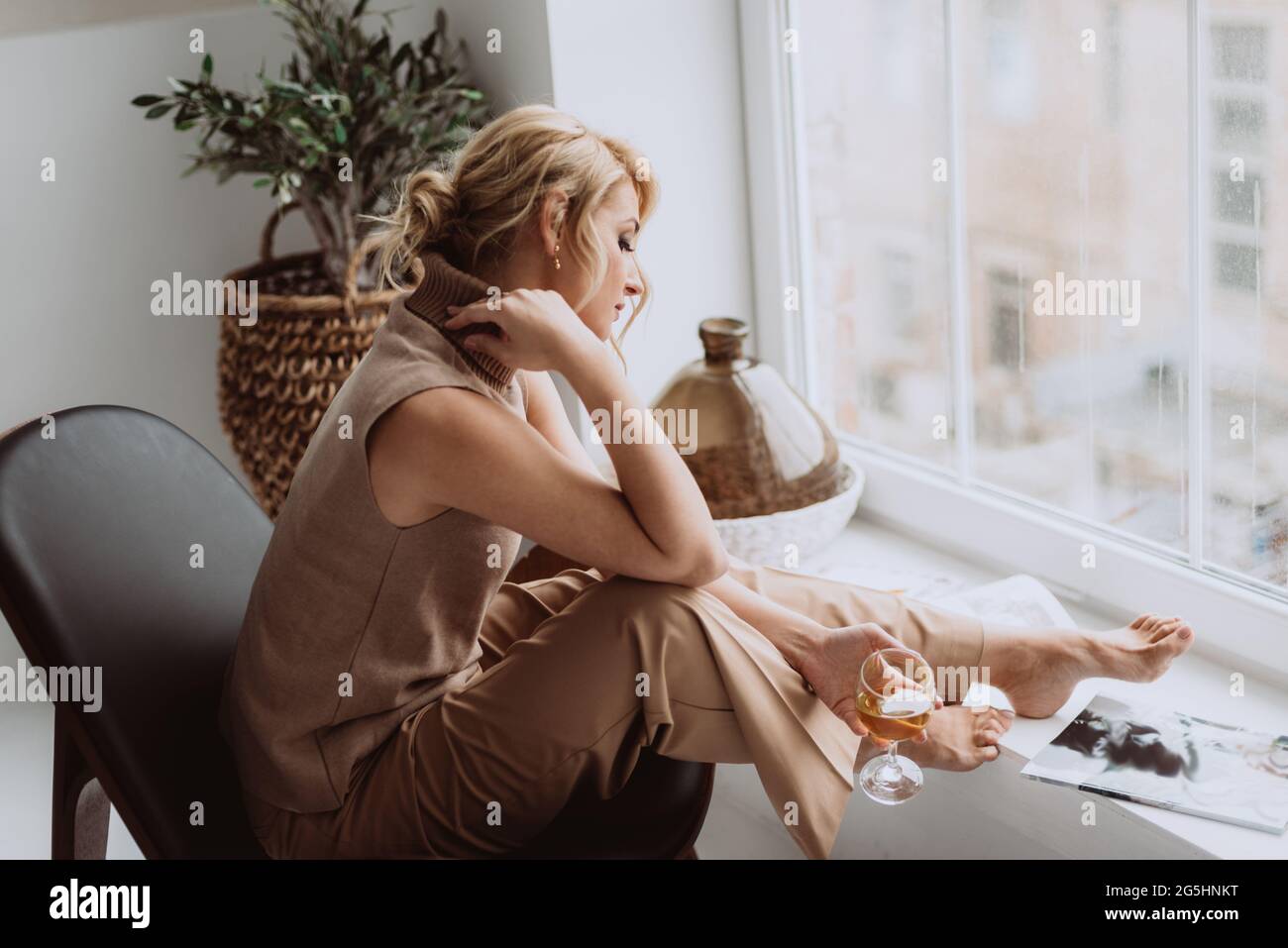 Das nachdenkliche, ruhige Modell hält ein Glas Weißwein und blickt aus dem Fenster. Schöne Frau in einem modernen Interieur. Weicher, selektiver Fokus. Stockfoto