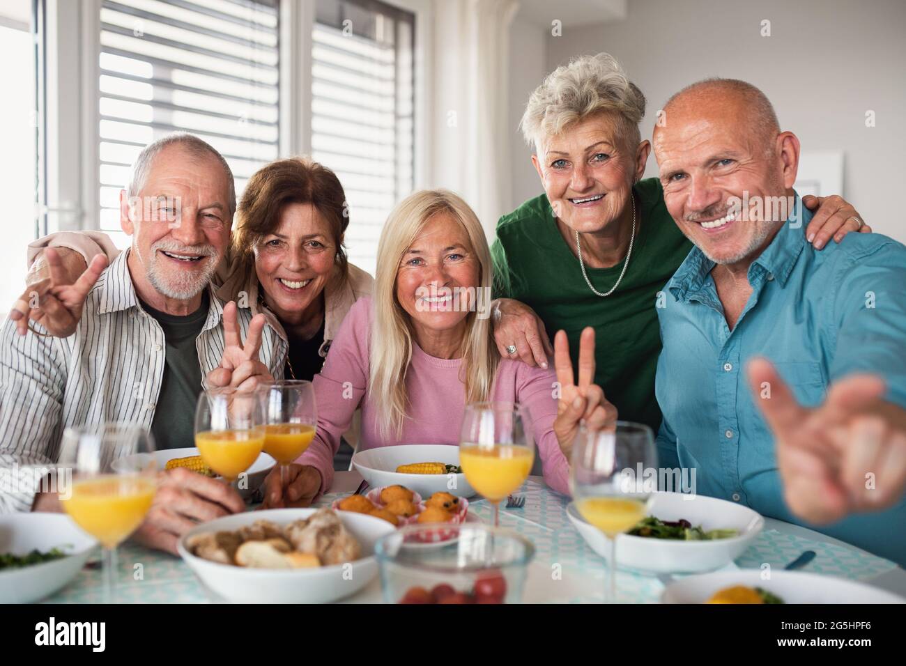 Gruppe älterer Freunde, die in einem Gebäude feiern und beim Essen auf die Kamera schauen. Stockfoto