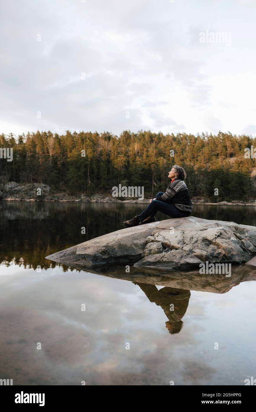 Weibliche Wanderin, die auf Felsen am See gegen den Himmel sitzt Stockfoto