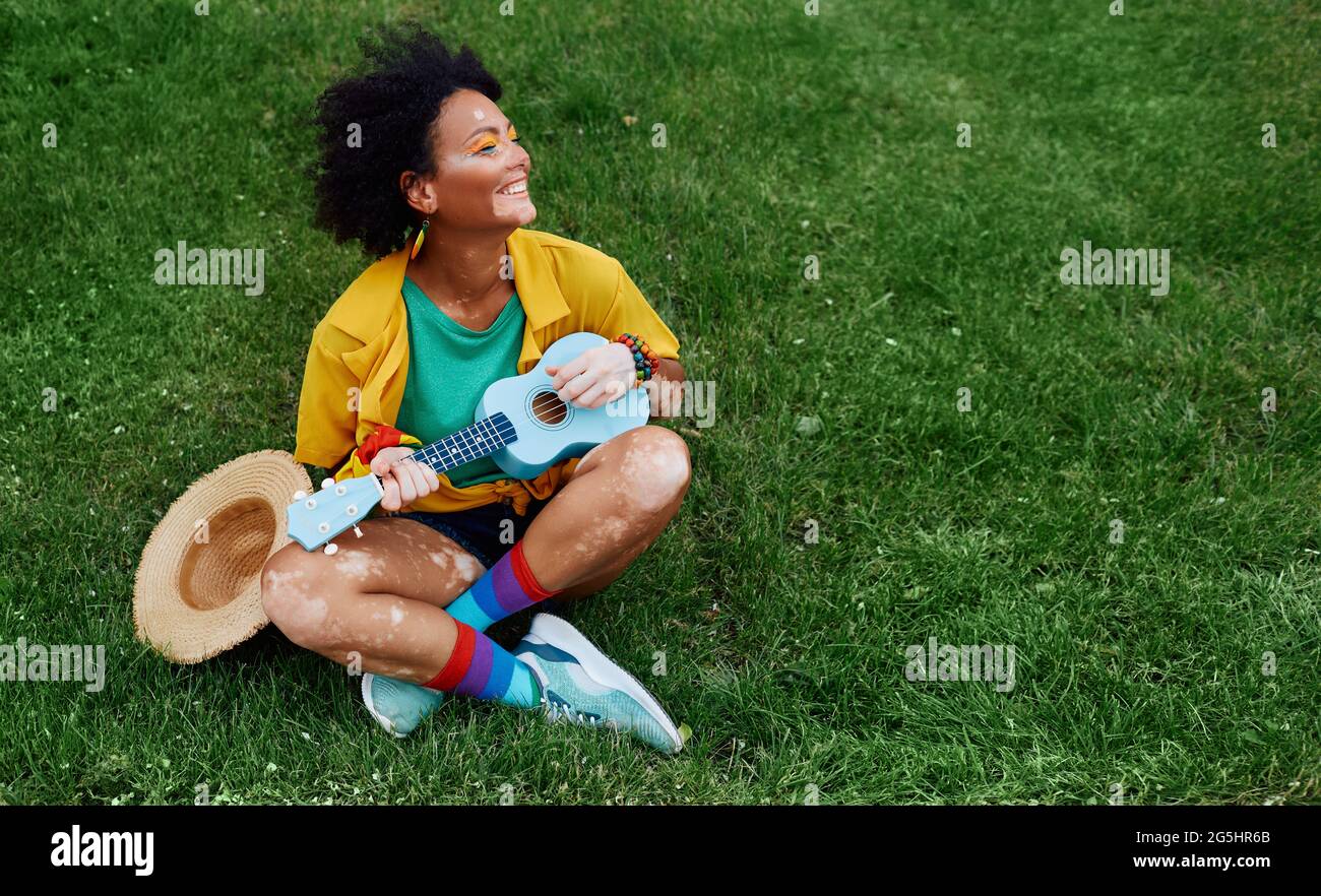 Rastafarian fun woman ist leidenschaftlich über das Spielen der Ukulele sitzen auf dem grünen Rasen, sie genießt Sommer und Musik Stockfoto