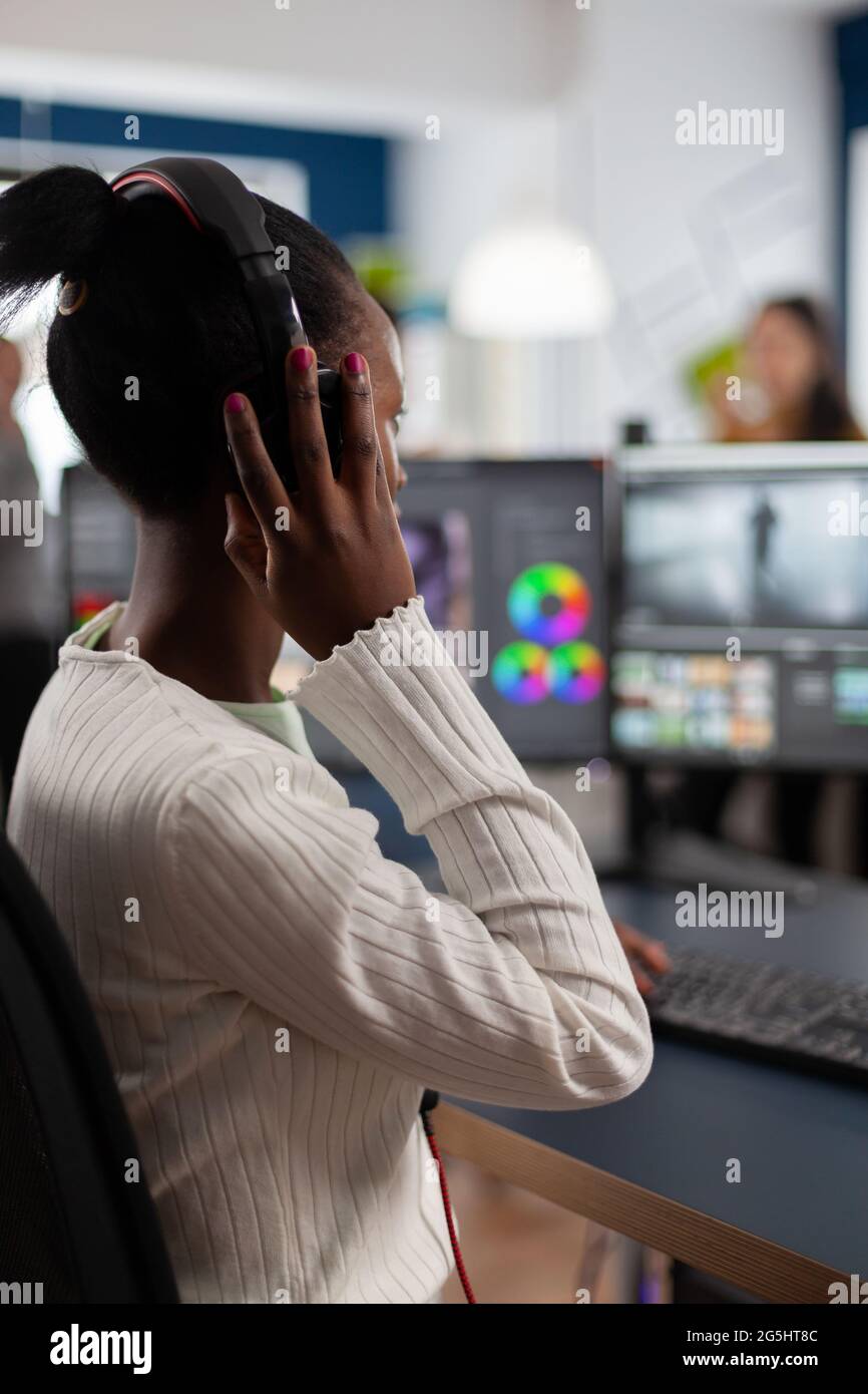 Schwarzer Videoeditor, der mit Filmmaterial auf dem pc mit zwei Monitoren arbeitet und Kopfhörer aufsetzt. Afrikanischer Videofilmer, der mit Post-Production-Software Filme bearbeitet, die in einem Büro der Kreativagentur arbeitet. Stockfoto