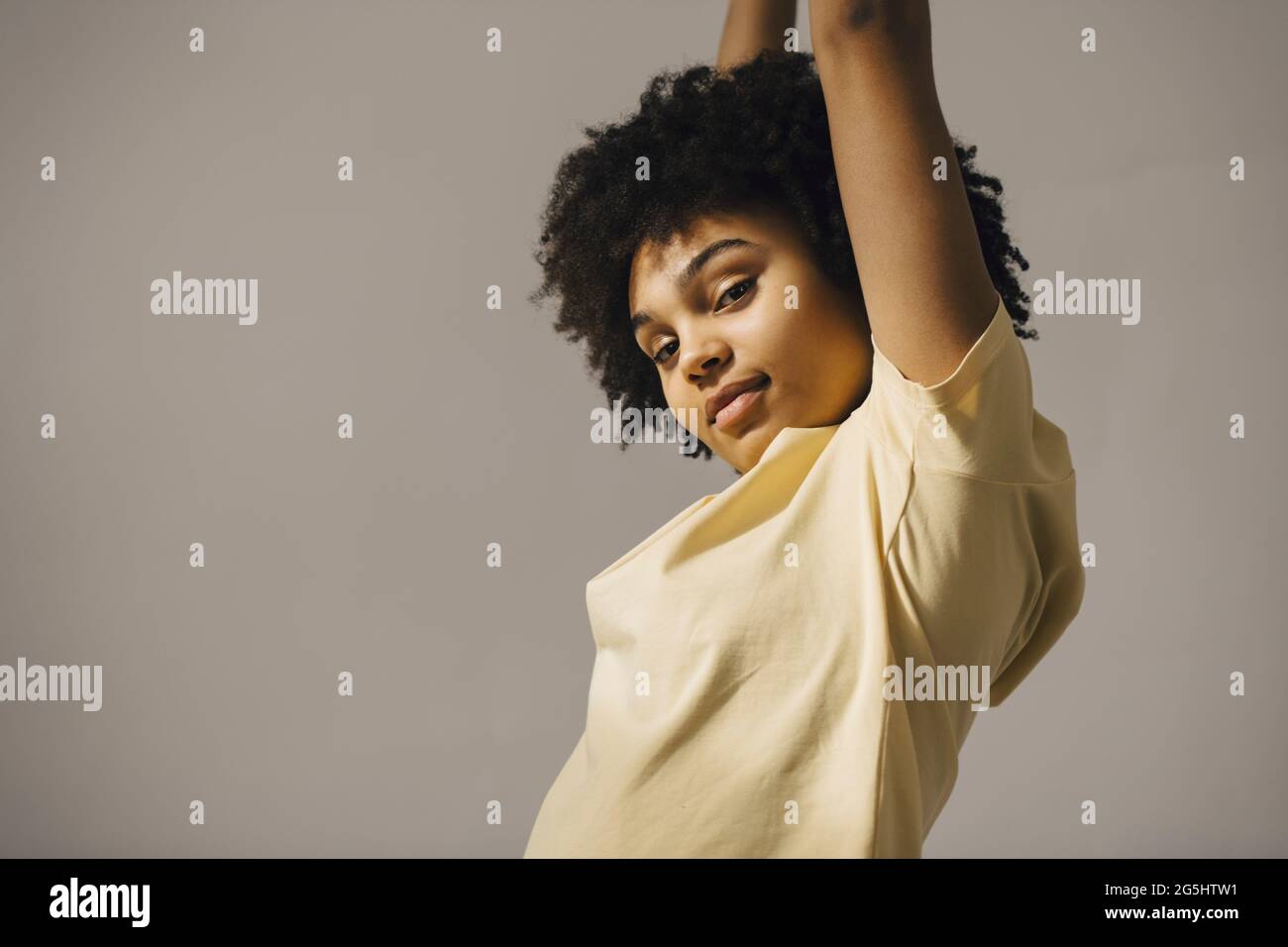 Porträt einer Frau mit lockigen Haaren, die die Arme vor beigem Hintergrund angehoben hat Stockfoto