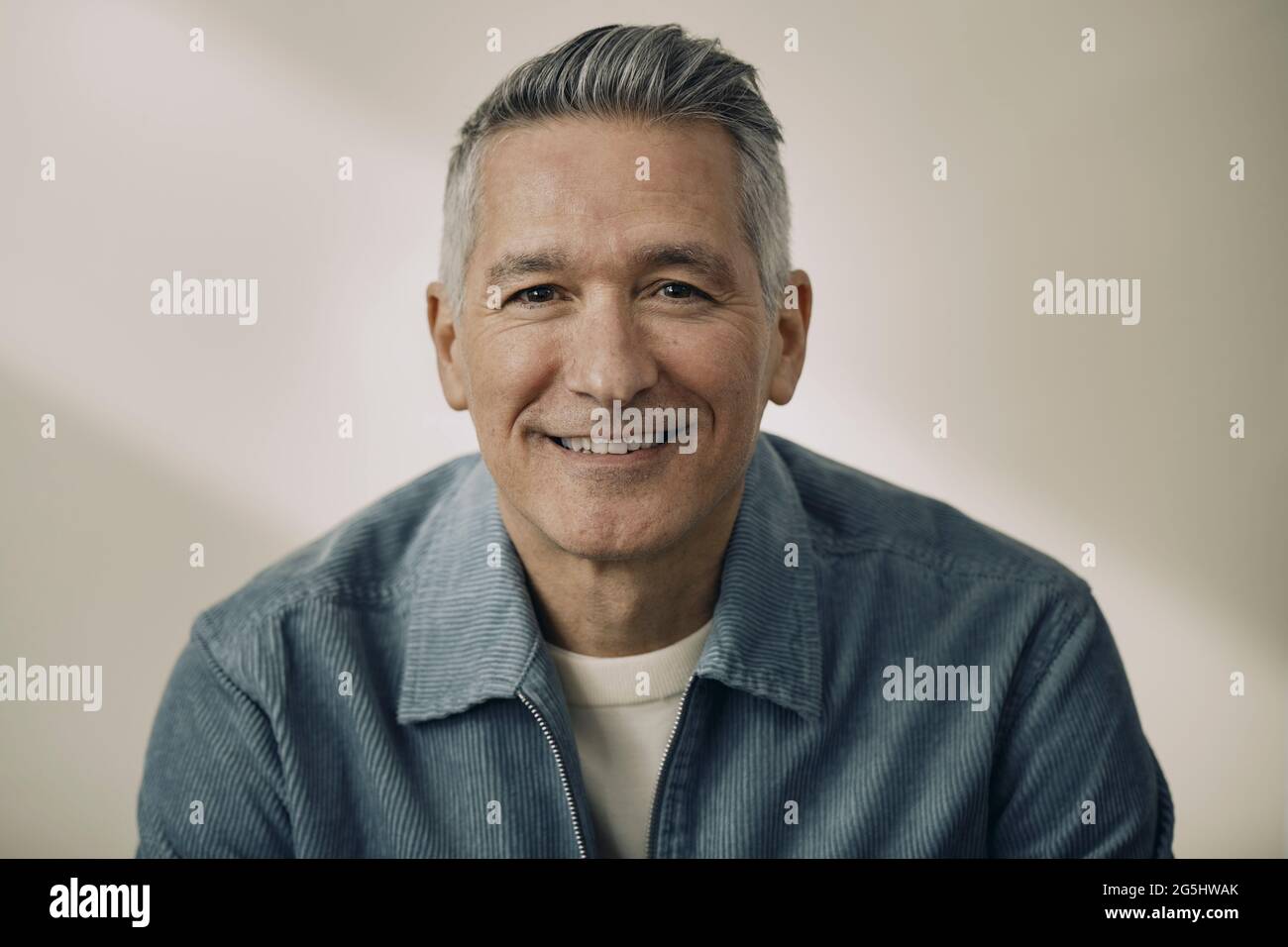 Porträt eines lächelnden, reifen Mannes, der eine blaue Jacke an der beigen Wand trägt Stockfoto