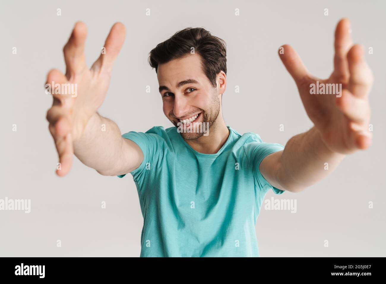 Fröhlicher gutaussehender Kerl lächelt und reicht die Hände zur Kamera isoliert über die weiße Wand Stockfoto