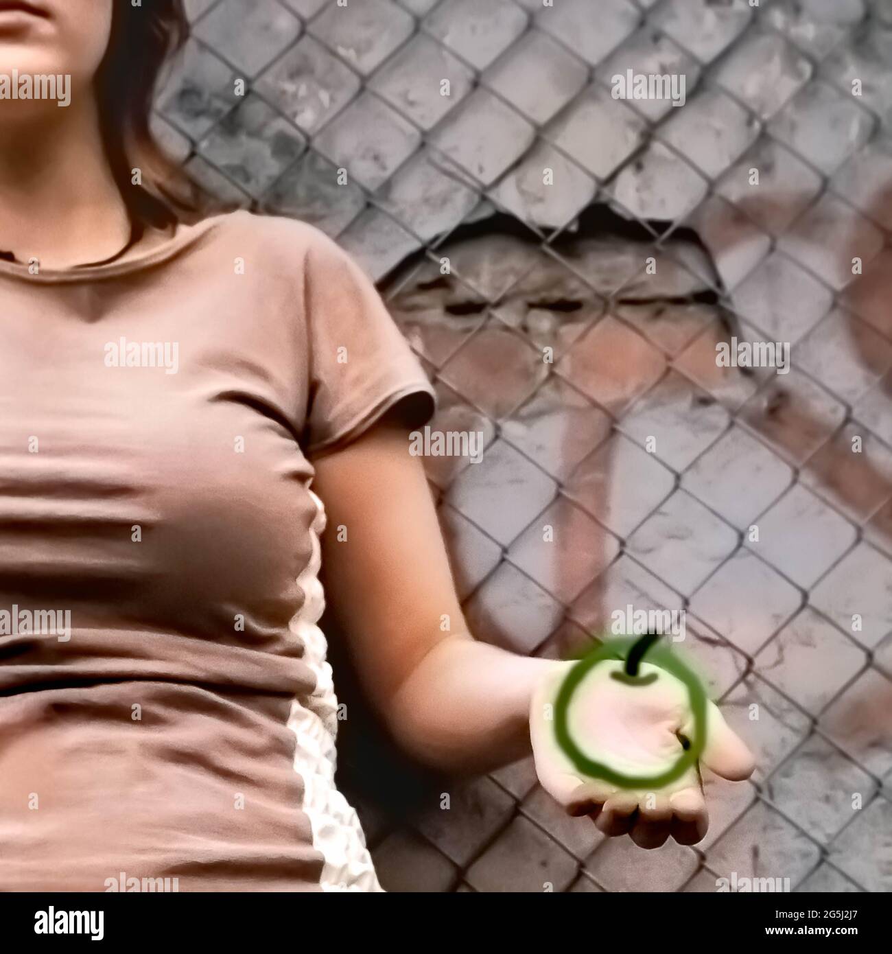 Ein junges kaukasisches, cooles Stadtmädchen, das einen grünen Apfel vor einer rauen, schäbigen Beton- und Ziegelmauer mit Graffiti und einem Gitterzaun hält Stockfoto