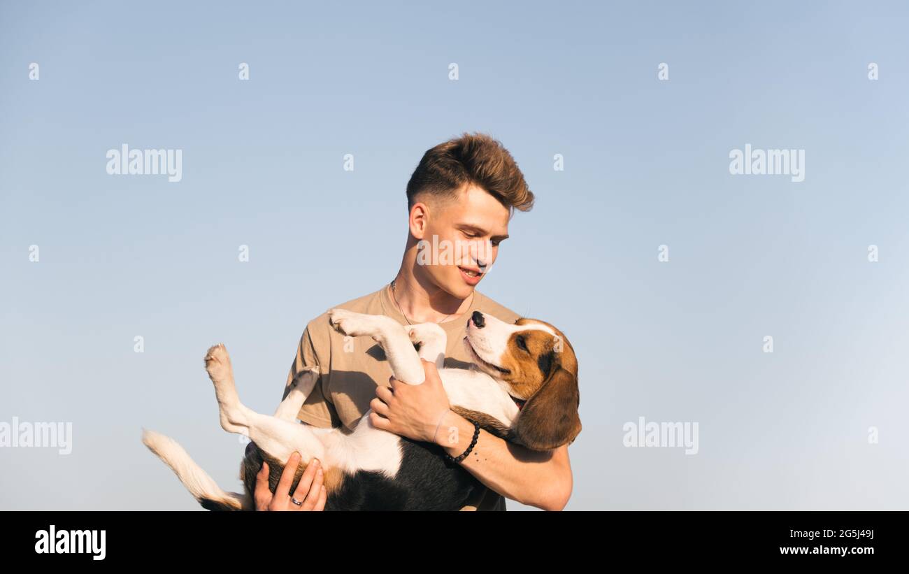 Der junge Mann hält seinen glücklichen Beagle-Hund in den Händen, der gegen den klaren blauen Himmel geschossen wurde. Lifestyle mit Haustieren, glücklicher Welpenbesitzer, reinrassiger Hund Stockfoto