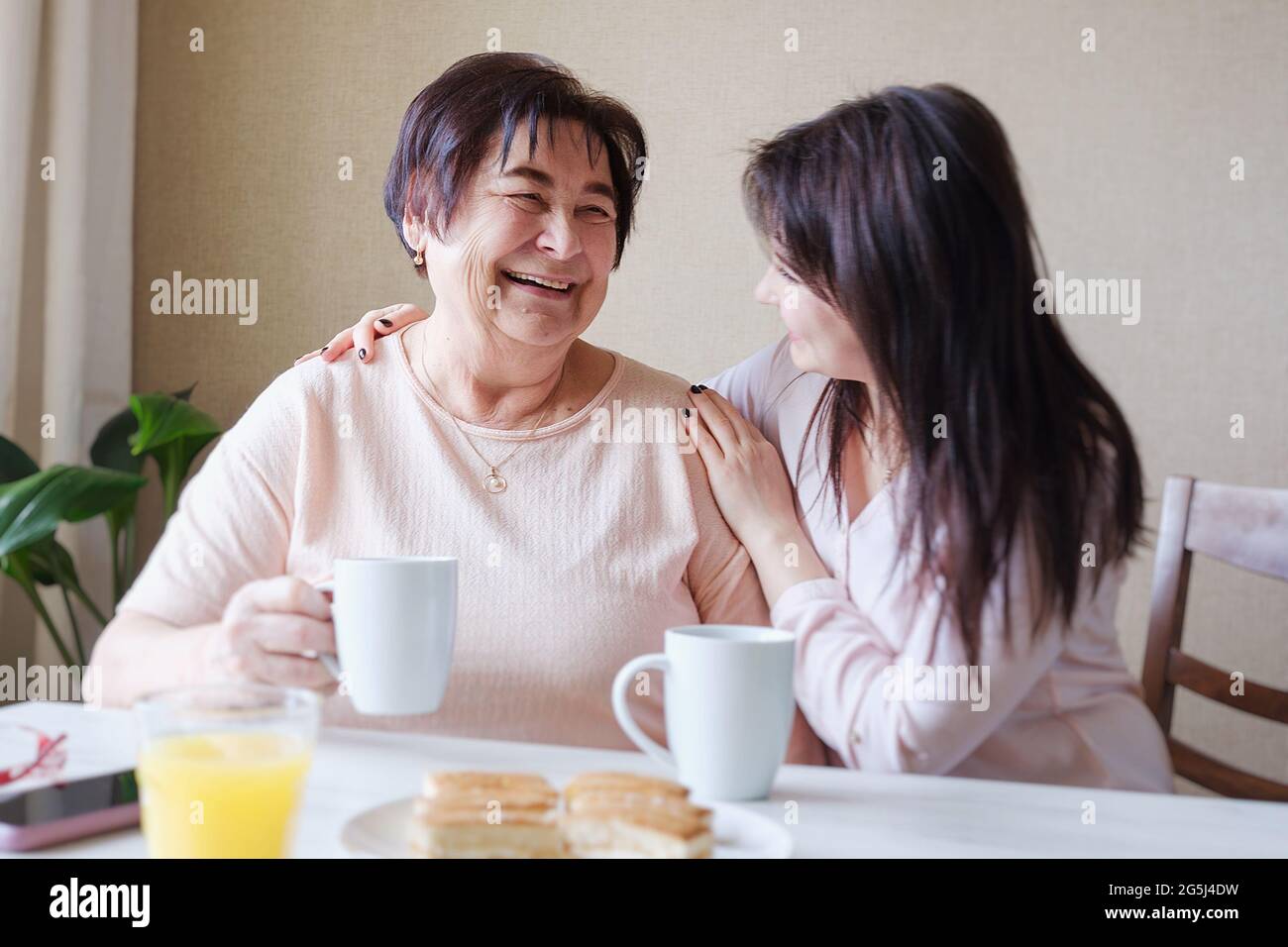 Glückliche junge Tochter umarmt eine ältere Mutter beim Frühstück am Tisch - zwei Generationen drücken Fürsorge und Wiedervereinigung aus - das Konzept o Stockfoto
