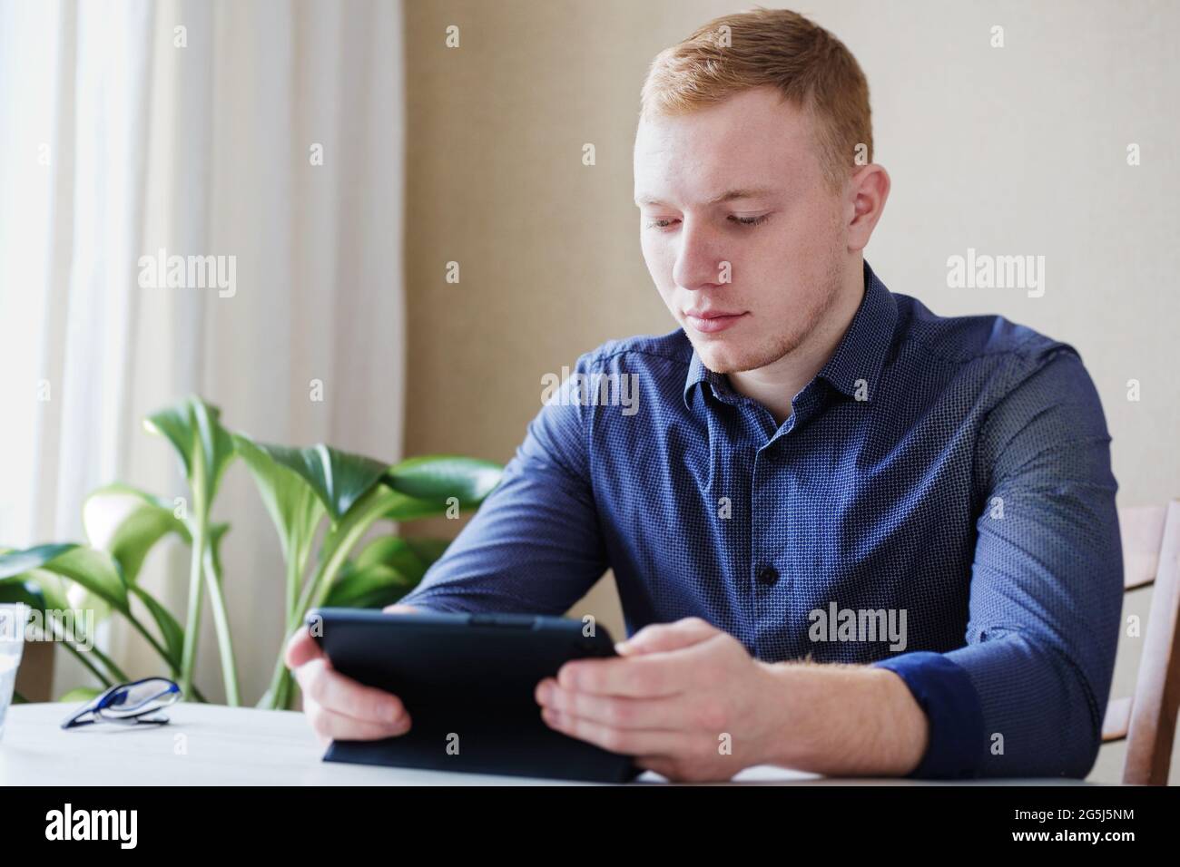 Junger Mann mit europäischem Auftritt liest die Nachrichten auf dem Tablet zu Hause - Konzept von Coworking und Menschen, die zu Hause arbeiten - Millennial Surfen im Inter Stockfoto