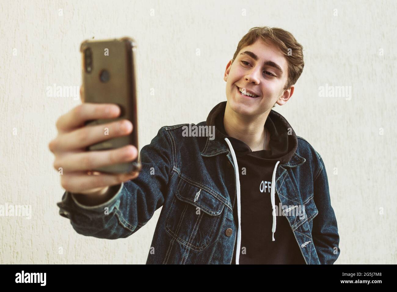 Ein Teenager macht ein Selfie mit seinem Handy auf einem hellen Hintergrund - ein glücklicher junger Mann macht ein Foto von sich selbst mit einem Telefon - EIN Teenager in einer Denim-jacke Stockfoto