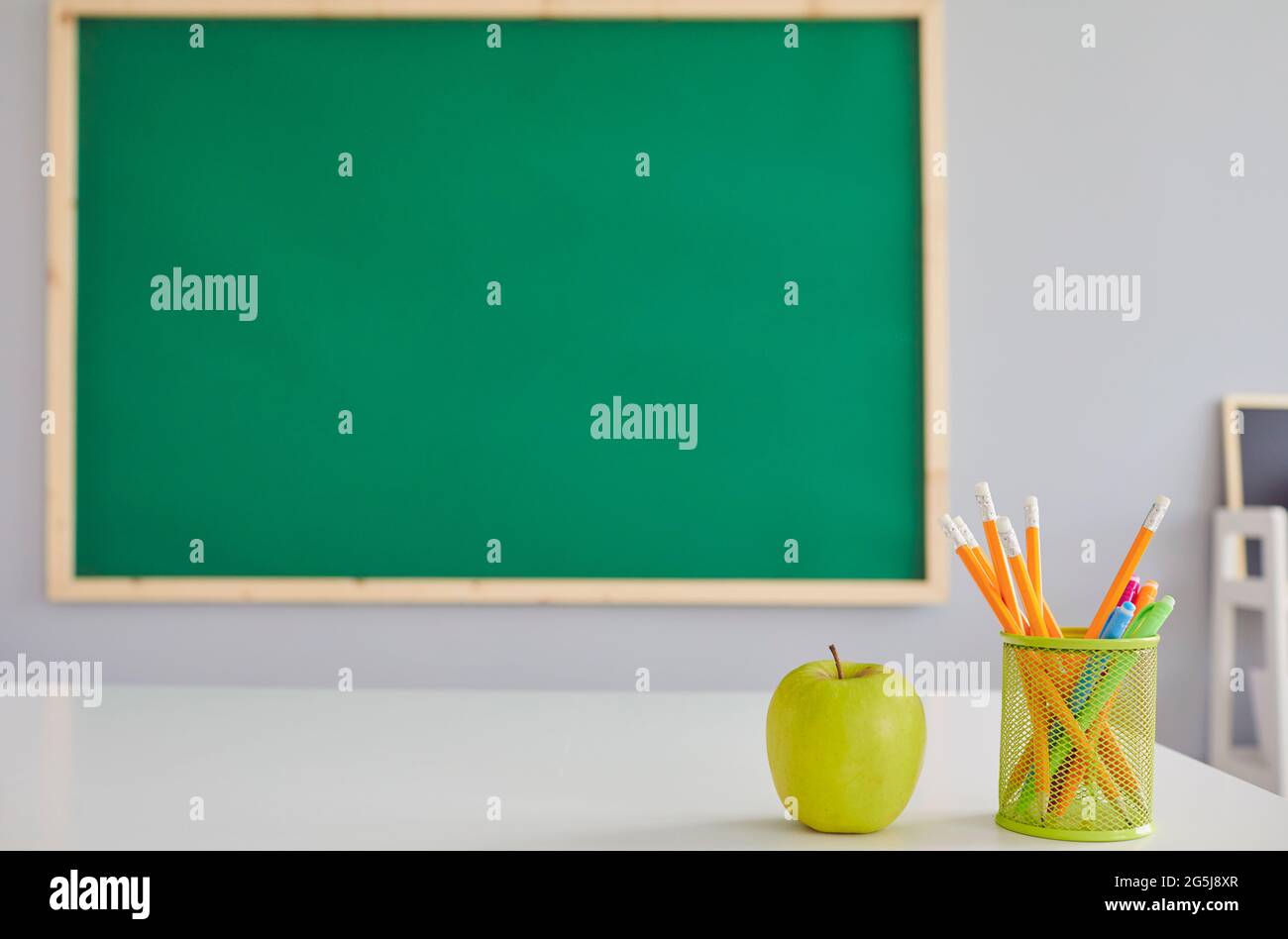 Grüner Apfel und Schreibwaren auf dem Schreibtisch des Lehrers im Klassenzimmer, leerer Raum. Obst und Schulbedarf auf dem Tisch Stockfoto