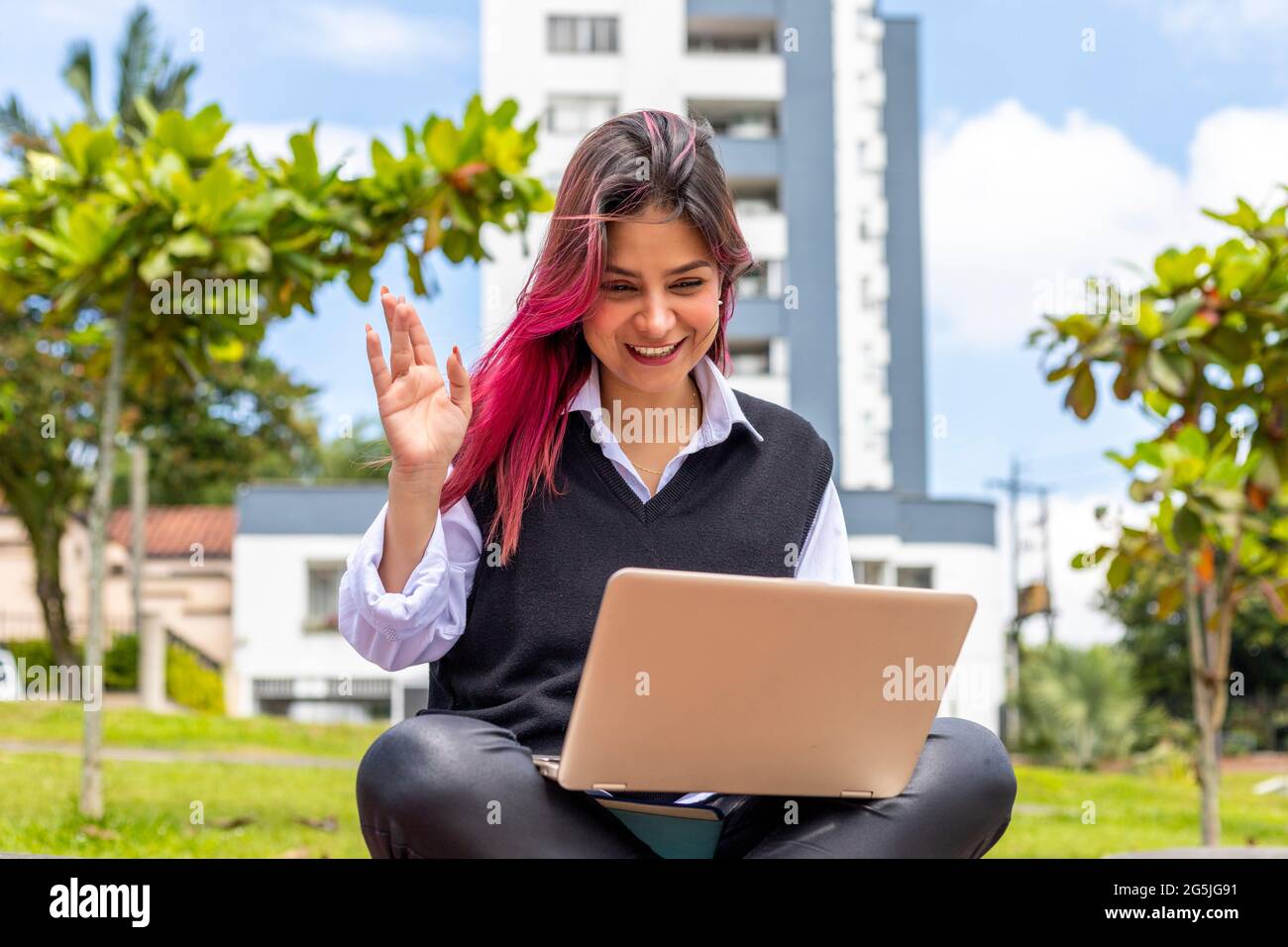 Junge Frau, die im Freien sitzt und auf dem Bildschirm ihres Laptops winkt Stockfoto