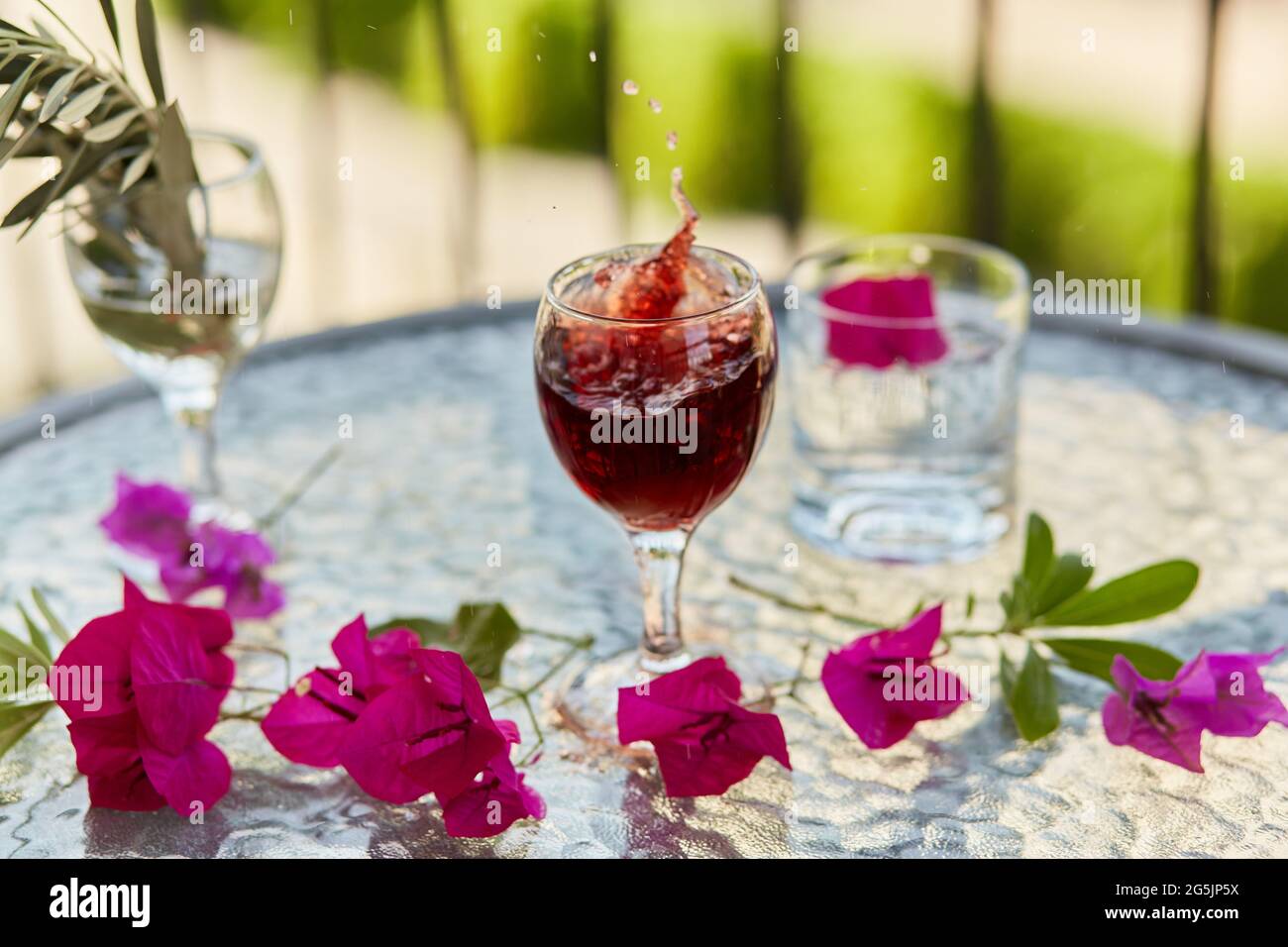 Explosion von eleganten und bunten Glas Rotwein, Wein spritzt im Glas. Sommercocktails im Hintergrund. Dekorative rosa Blüten und eine Spanke Stockfoto