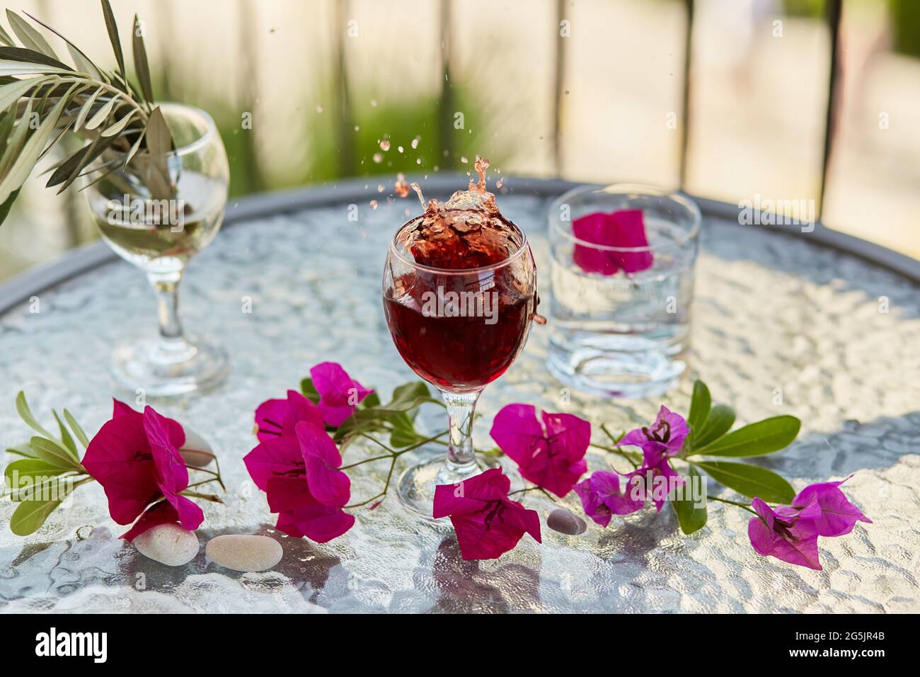 Elegante und farbenfrohe Explosion von Rotwein. Sommercocktails im Hintergrund. Dekorative rosa Blumen und der Zweig des Olivenbaums im Glas. Glyp Stockfoto