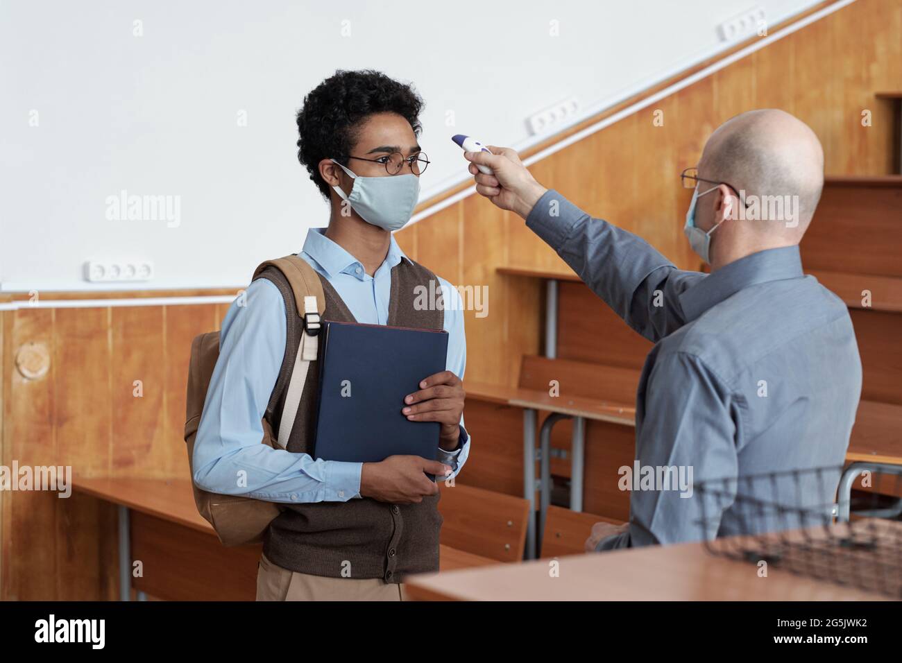 Lehrer, der die Körpertemperatur des Schülers in einer Maske im Klassenzimmer misst Stockfoto