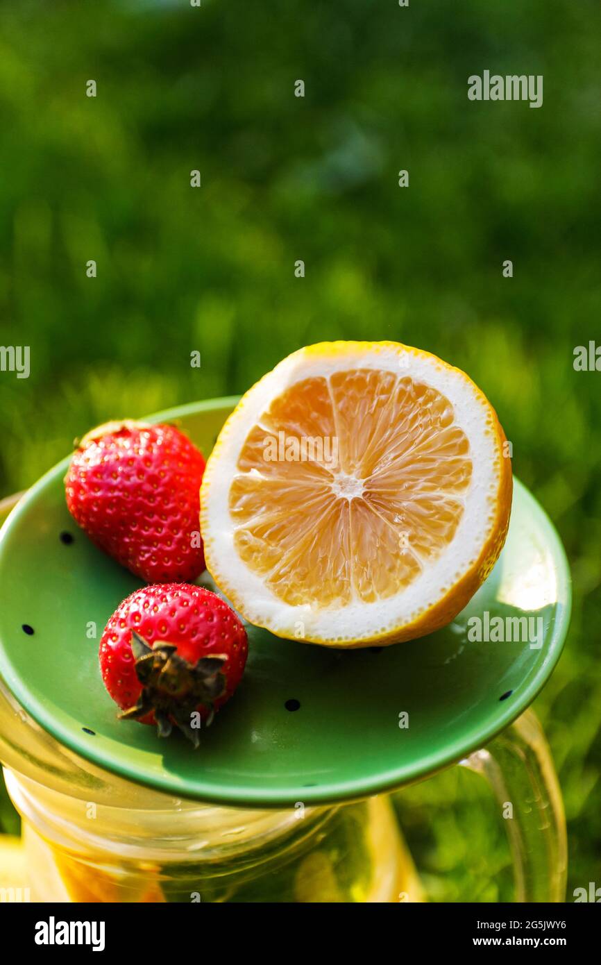 Unschärfe-Nahaufnahme-Untertasse mit Zitrone und zwei Erdbeeren stehen auf Glaskrug mit Limonade. Verschwommener Grashintergrund. Sommerfrüchte draußen, draußen Stockfoto