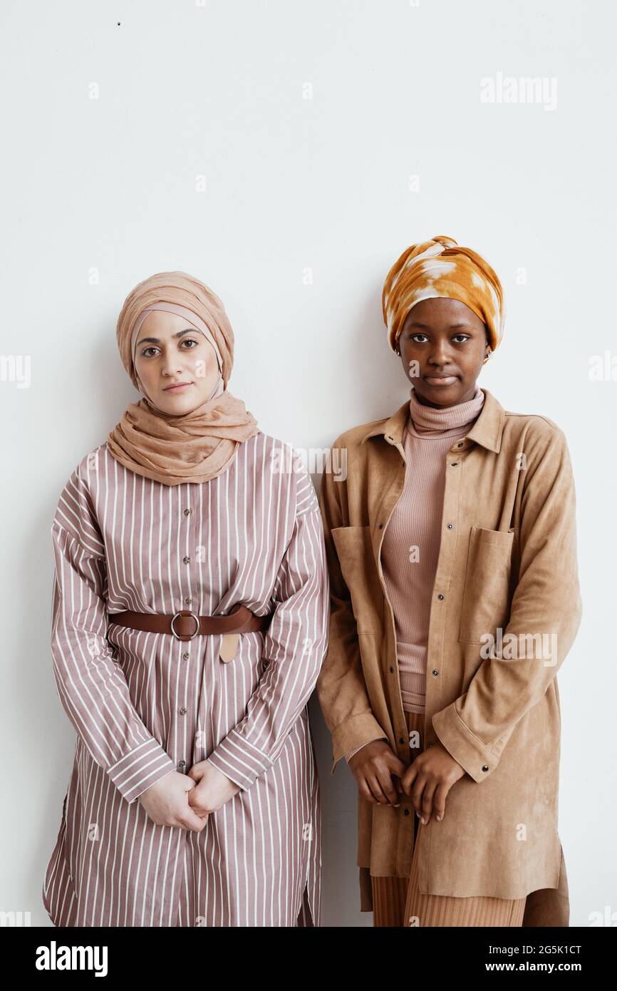 Minimales vertikales Porträt zweier ethnischer junger Frauen, die auf die Kamera schauen, während sie gegen eine weiße Wand stehen Stockfoto