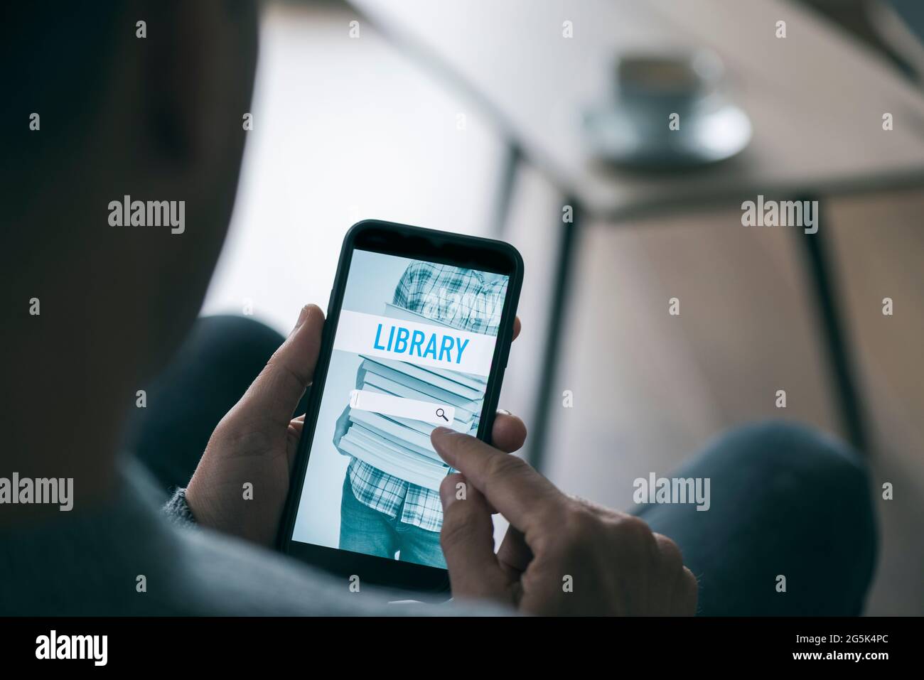 Ein junger Kaukasusmann, der an einem Couchtisch in seinem Wohnzimmer sitzt, sucht mit seinem Smartphone ein eBook, das er mit einer Simulae in einer Online-Bibliothek ausleihen kann Stockfoto